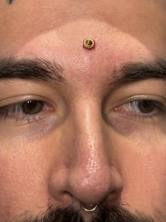 Close-up of a person's face with a decorative forehead piercing and a septum piercing, eyes looking downward.