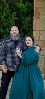 A man with a beard and a woman with purple hair stand close together outdoors, smiling, with trees and a brick wall behind them.