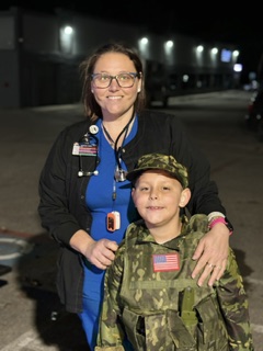 A woman in medical scrubs and a black jacket stands with a young boy in camouflage military uniform outdoors at night, smiling.