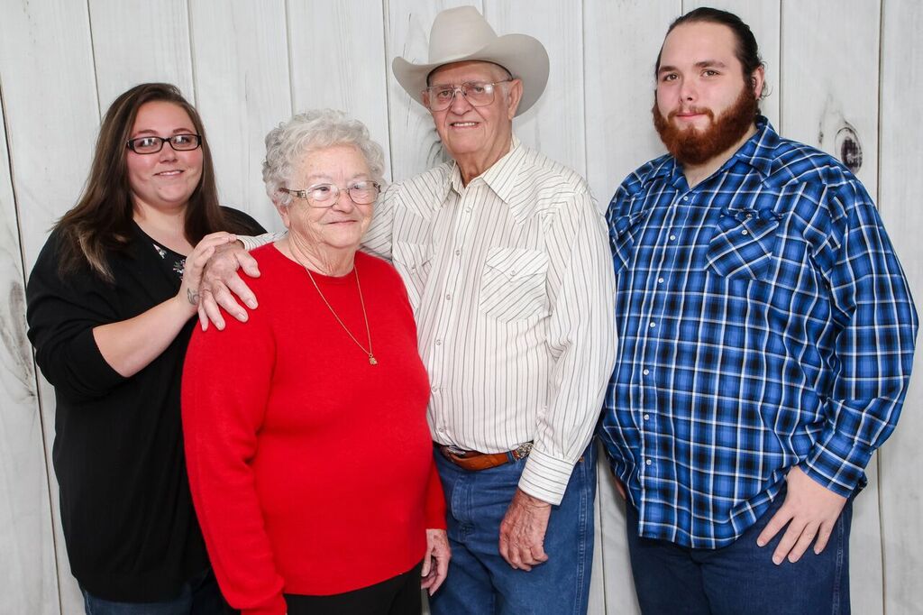 Four people standing together in front of a white wooden wall, smiling, with one person wearing a cowboy hat.