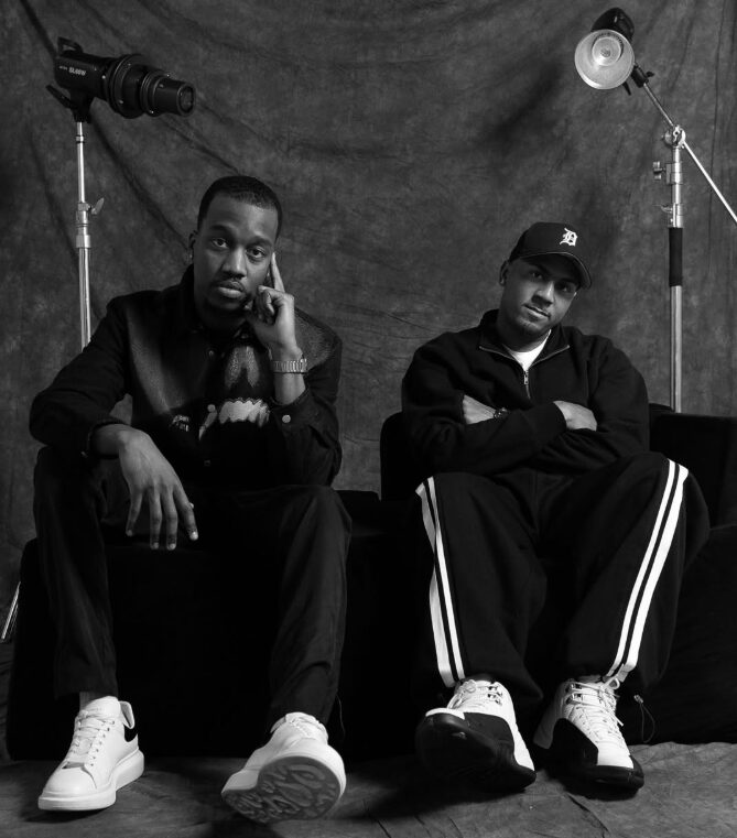 Two men sit on a black couch in a photography studio with lighting equipment, one resting his head on his hand, the other crossing arms.