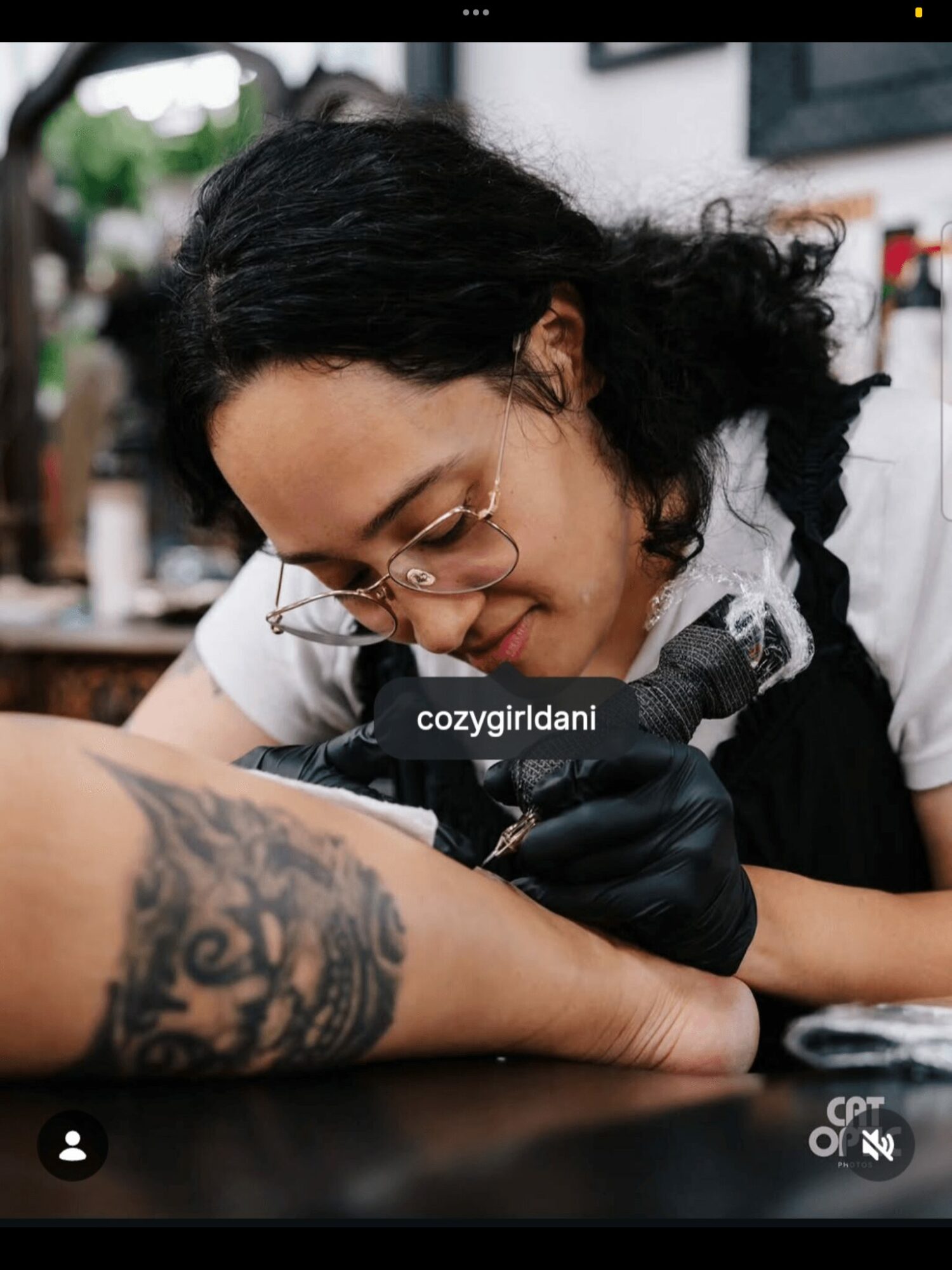 Tattooed arm being tattooed by a person with glasses and black gloves, in a tattoo studio.