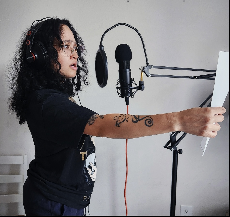 Young woman with curly hair, headphones, and glasses, pointing at a paper in front of a microphone setup.