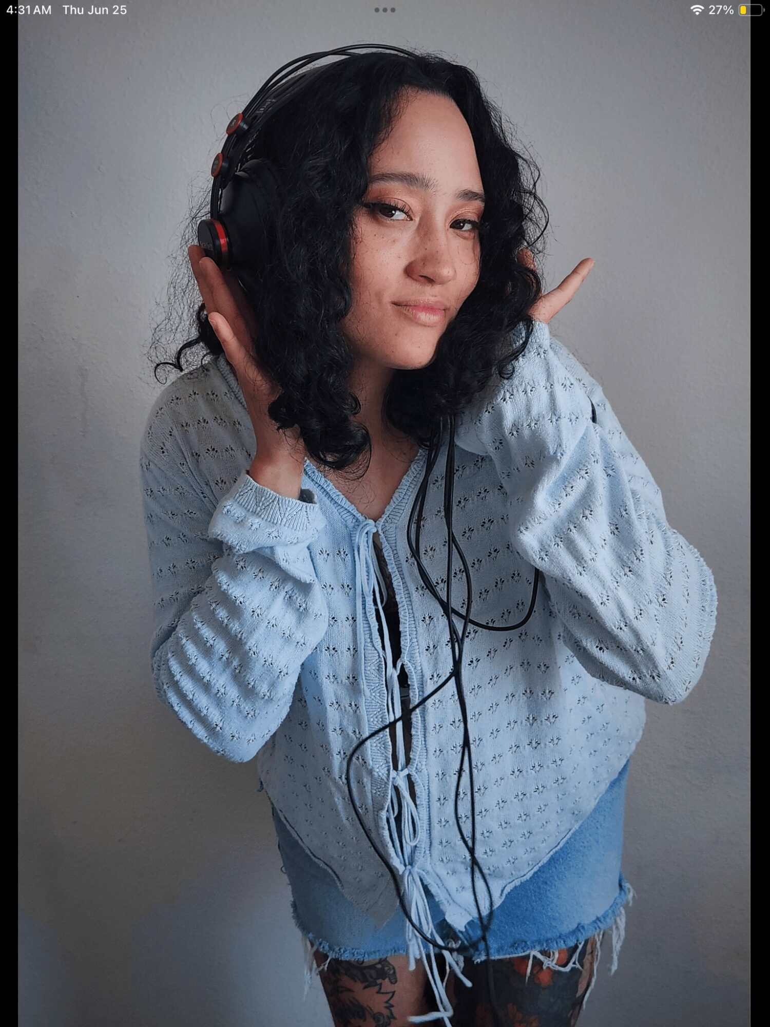 Young woman with curly hair wearing headphones, holding them with both hands, looking at the camera, gray background.