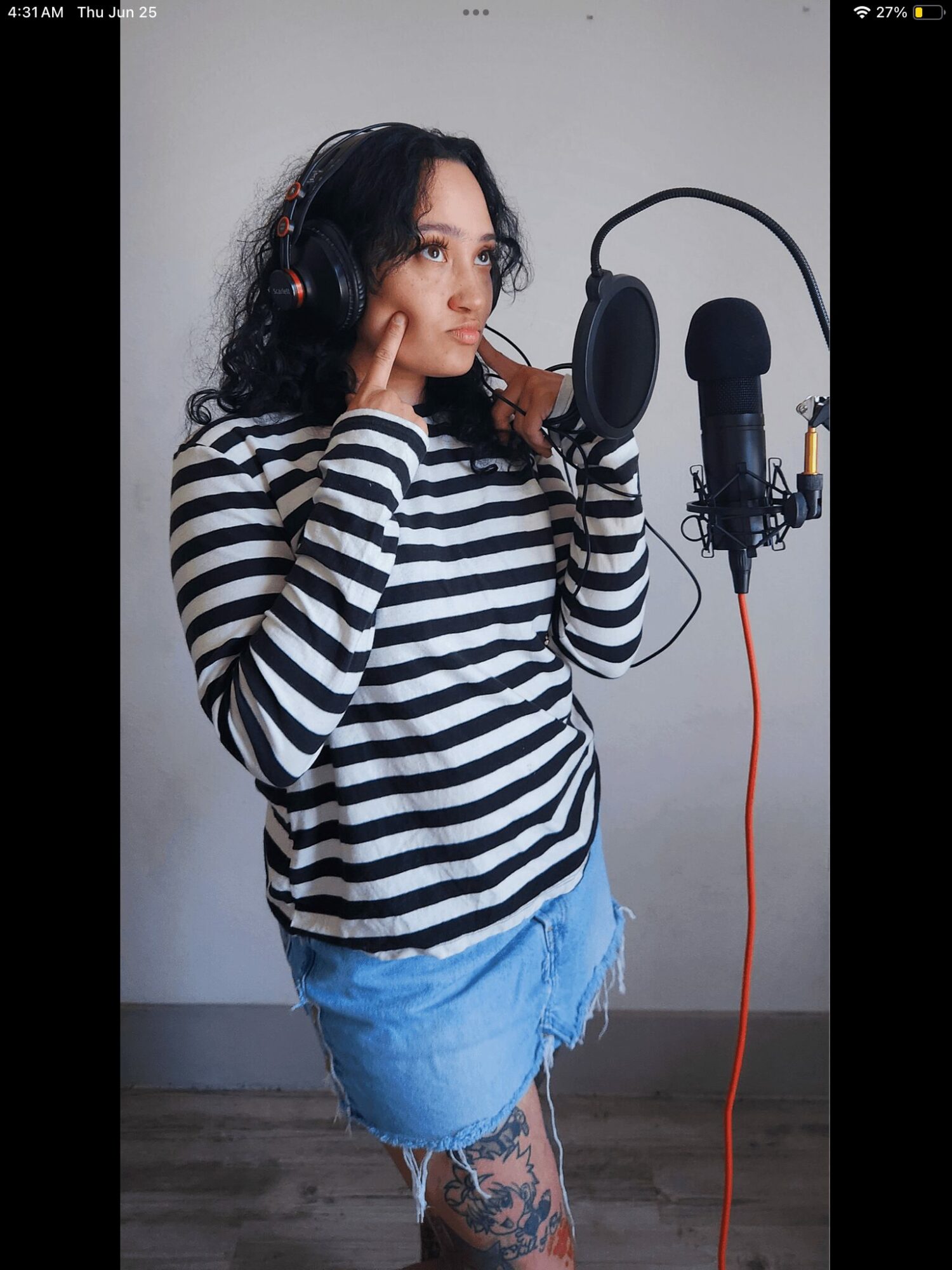 Young woman with headphones in a recording studio, wearing a striped shirt and denim skirt, standing near a microphone.