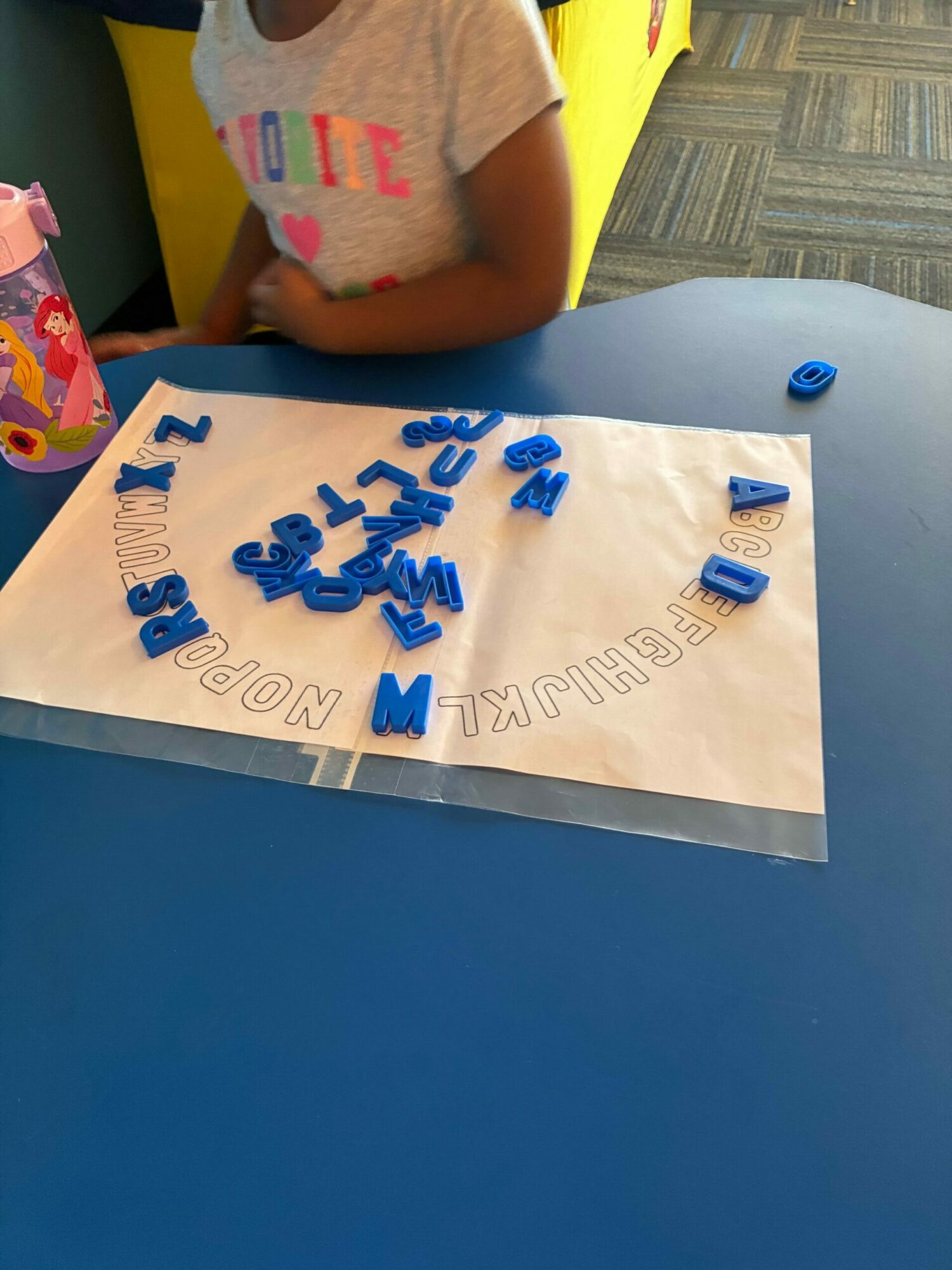 Child's arm and part of a yellow table with a poster displaying blue letters and numbers, on a blue wall.