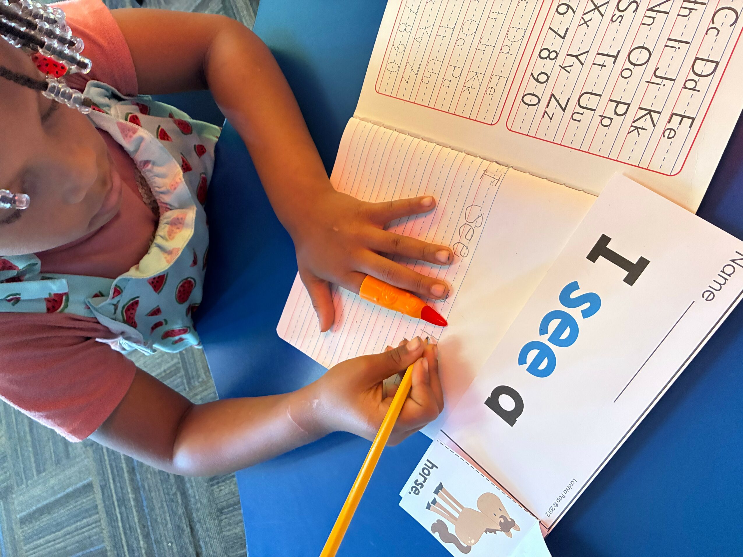 Child writing in a notebook with a yellow pencil, with an open book showing large letters and numbers, on a blue surface.