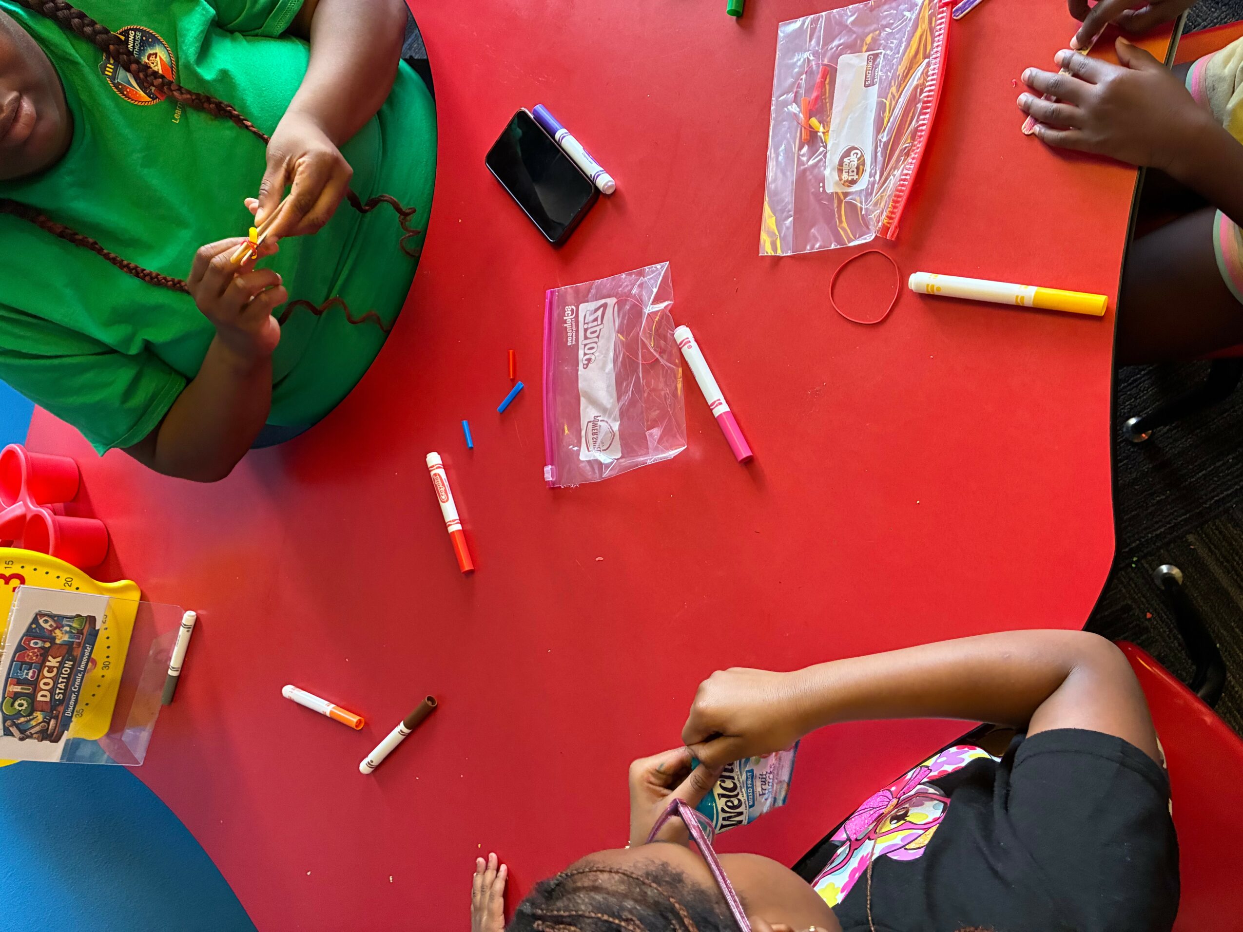 Children sitting around a red table with markers, a small black object, and plastic bags, engaging in a craft activity.