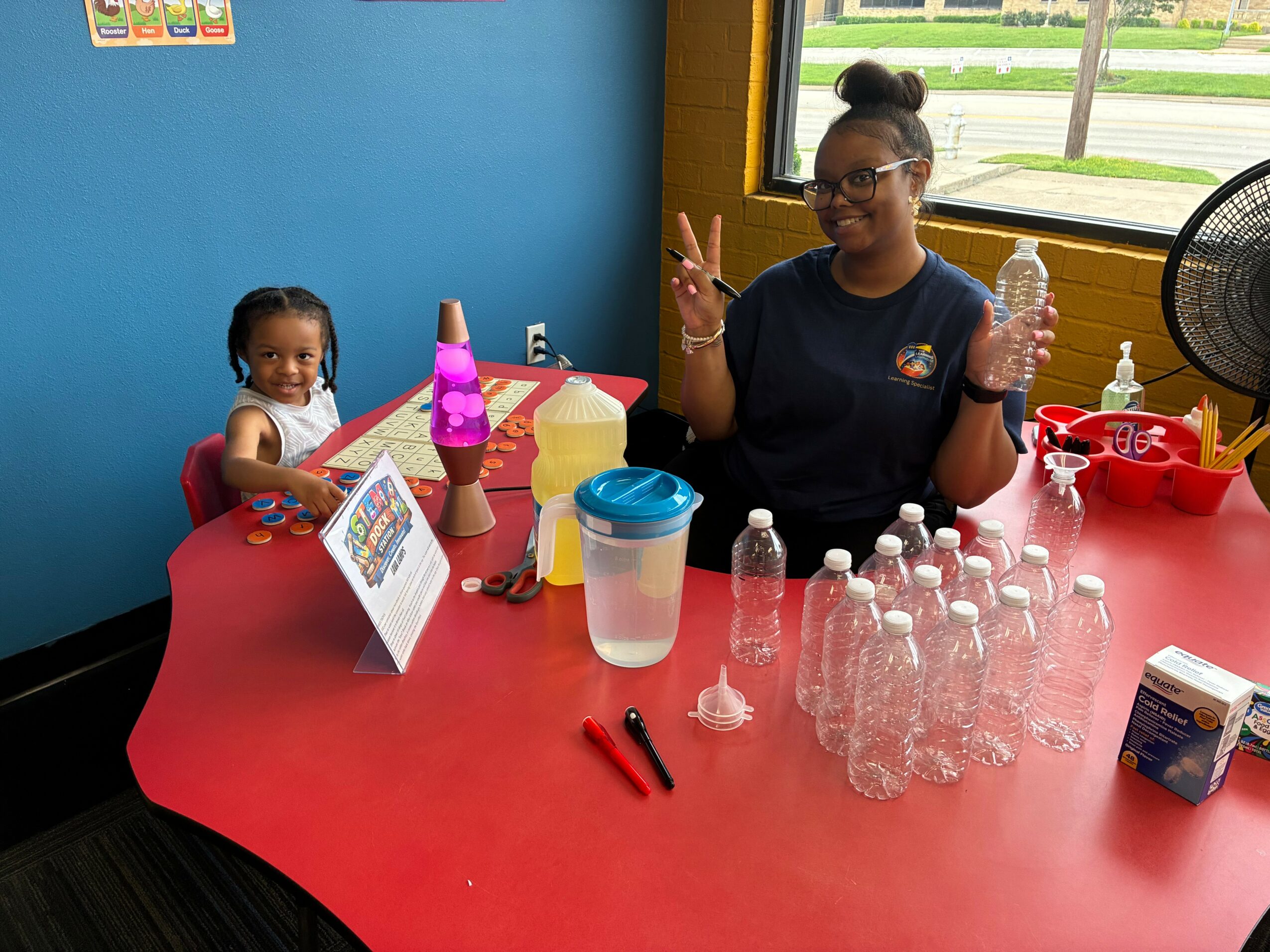 A woman and a young girl sit at a red table with water bottles, a sign, and cups in a room with blue walls and a window.