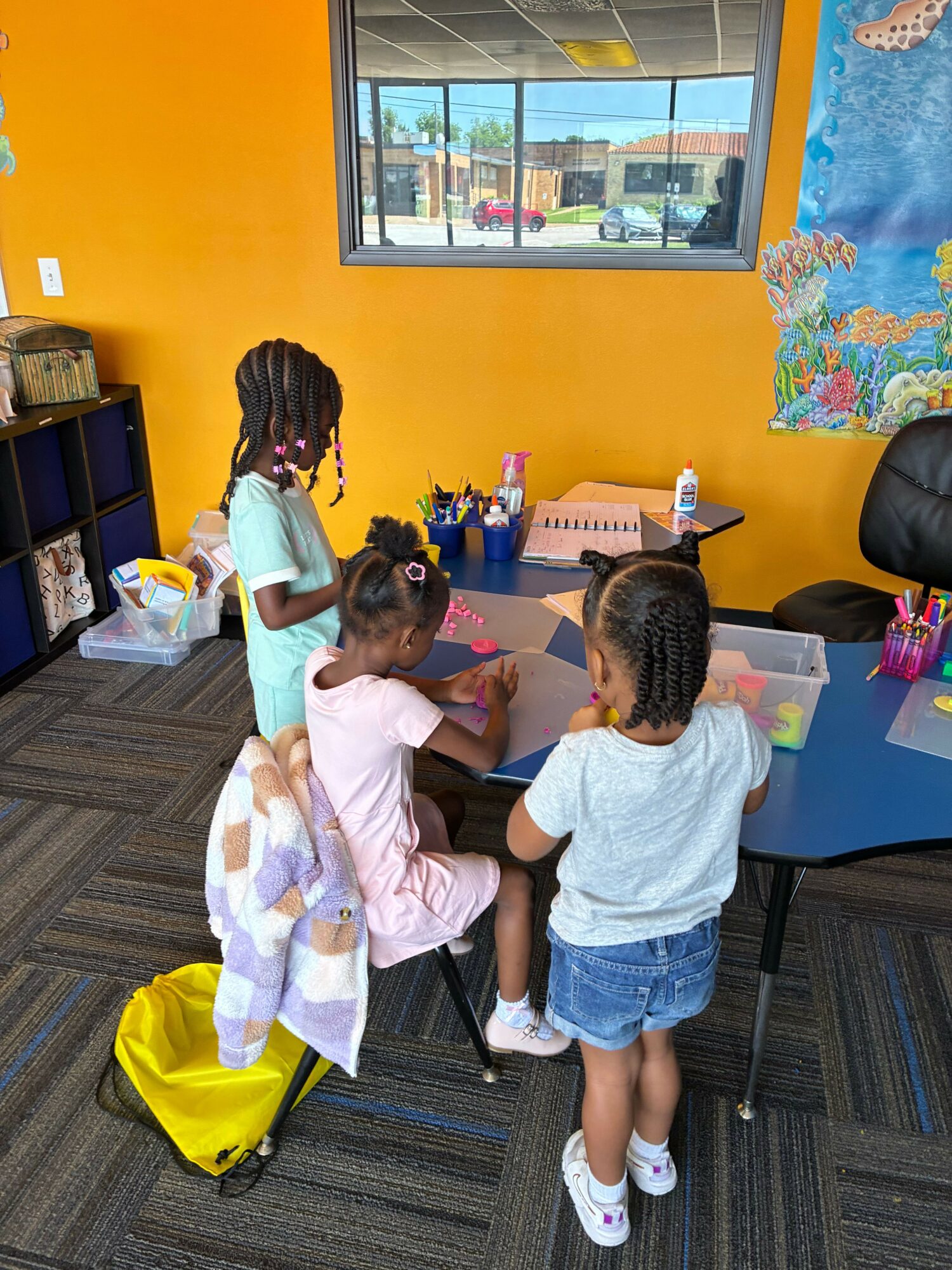Three children in a classroom near a window, engaging in an activity at a table with colorful walls and supplies.