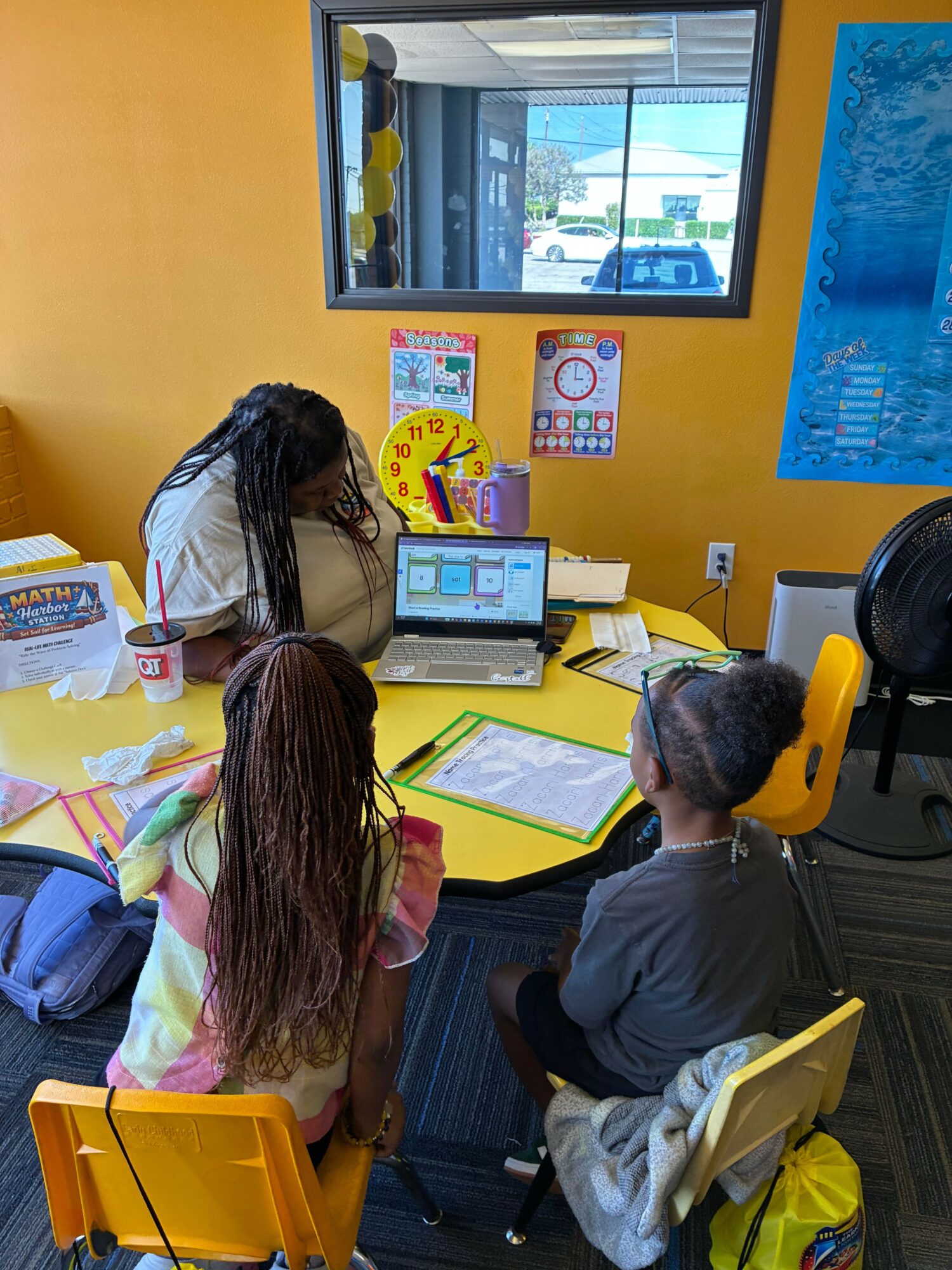 Two students work at a yellow table with a computer and papers in a classroom with yellow walls.