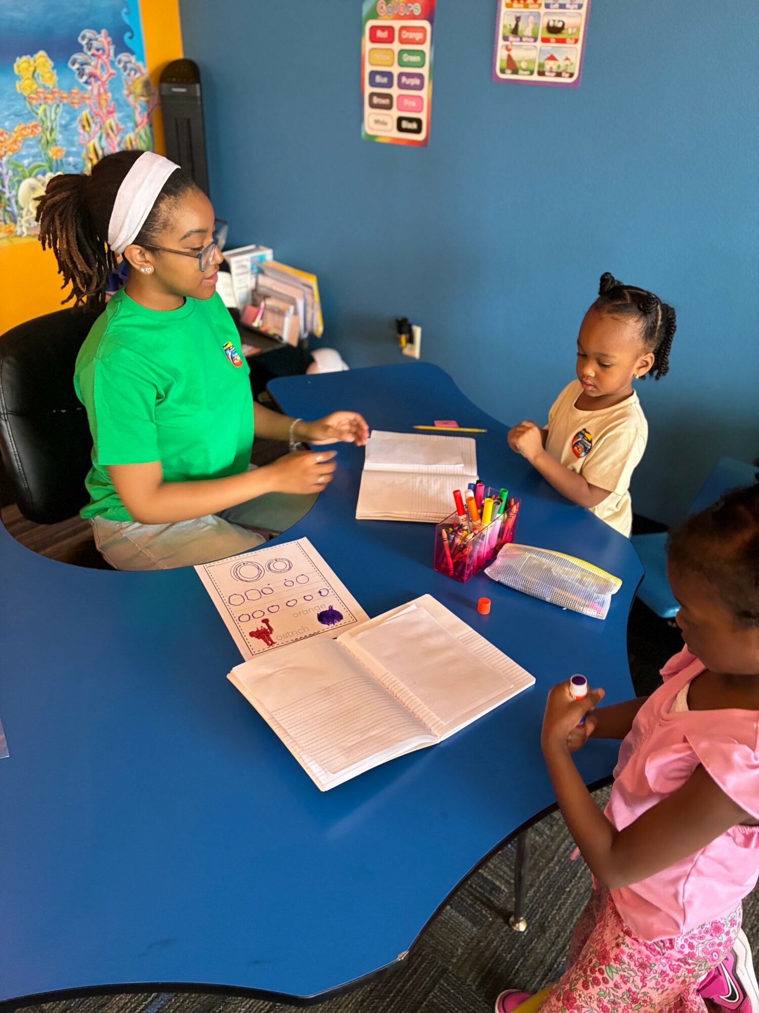 Adult woman and two children at a blue table in a classroom, engaging in learning activities with books and supplies.
