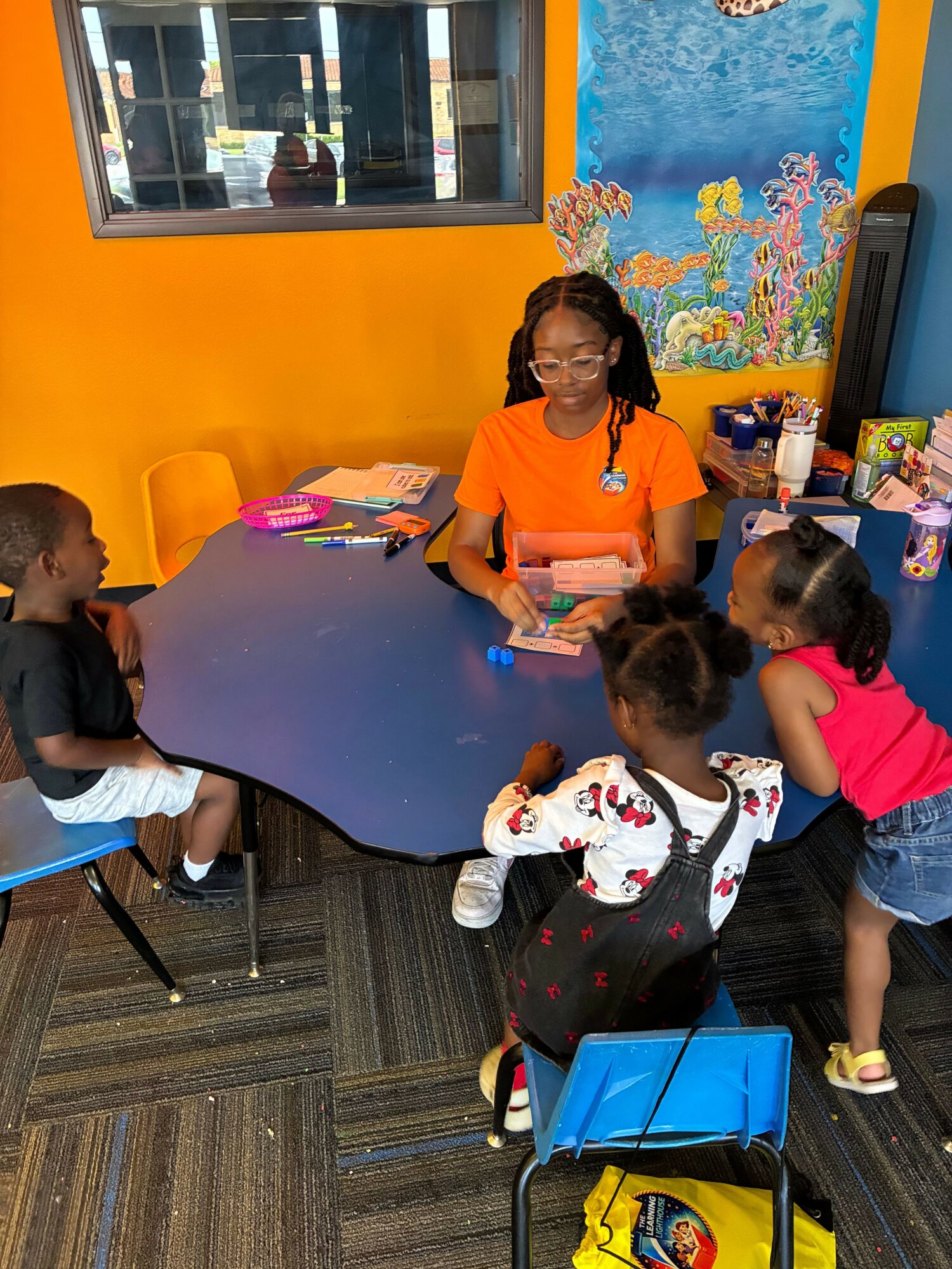 Children and an adult gather around a table in a colorful room, engaging in an activity. Bright yellow wall and window are visible.