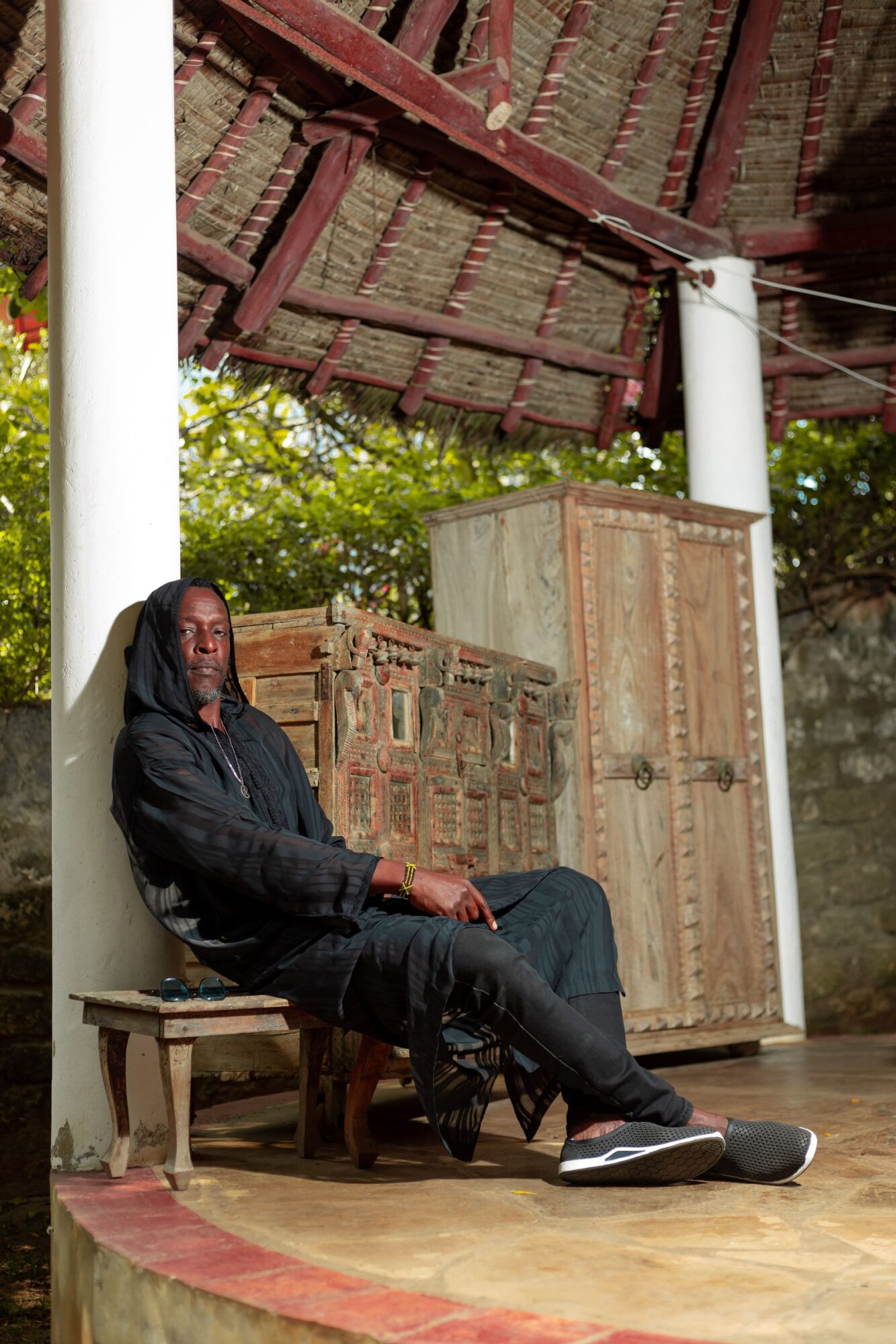 Person sitting on a bench under a thatched roof, with wooden furniture behind, outdoors with trees visible.