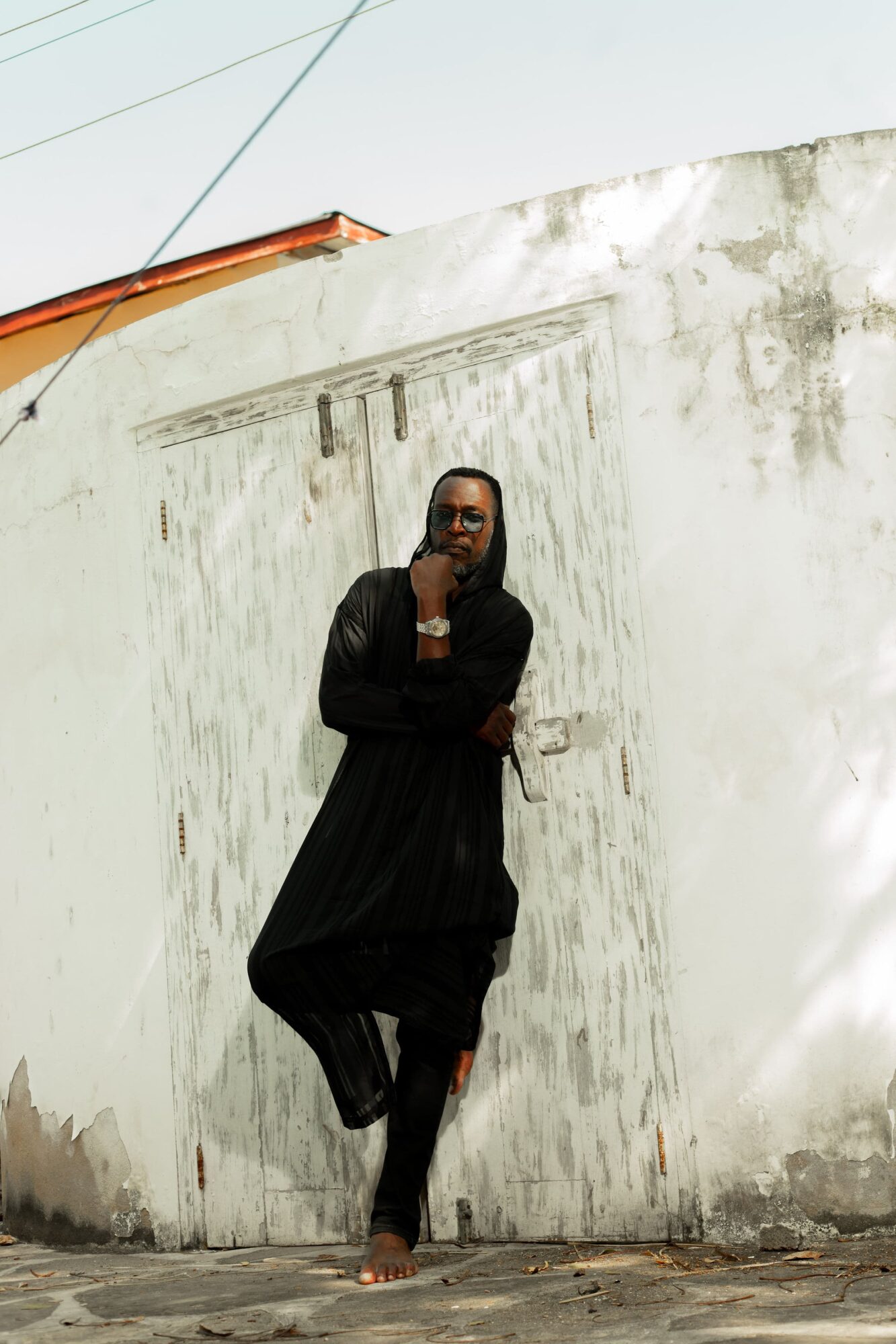 Person standing barefoot against a weathered white wall, wearing a black outfit, with arms crossed and a thoughtful expression.