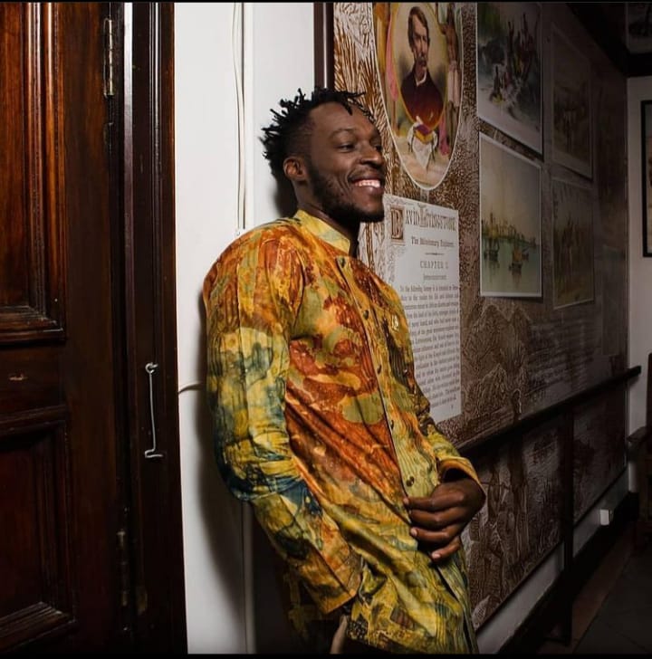 Person leaning against wall, smiling, wearing colorful patterned shirt, in indoor setting with framed pictures on wall.
