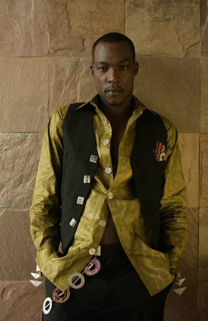 Young man with hands in pockets, wearing a yellow shirt and black vest, standing against a stone wall.