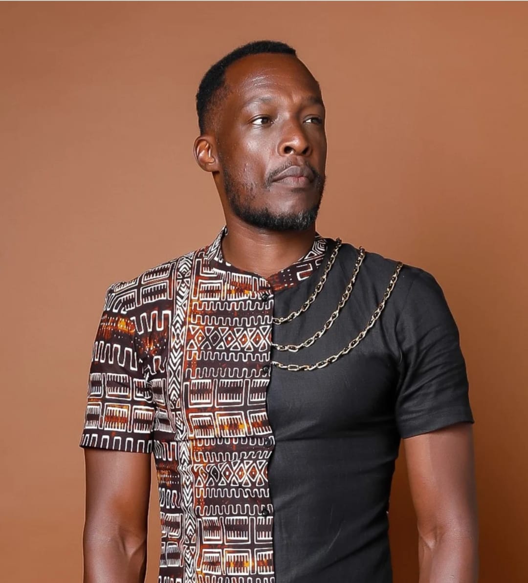 Man with short hair and beard wearing a black shirt with patterned and chain details, looking to the side, neutral background.