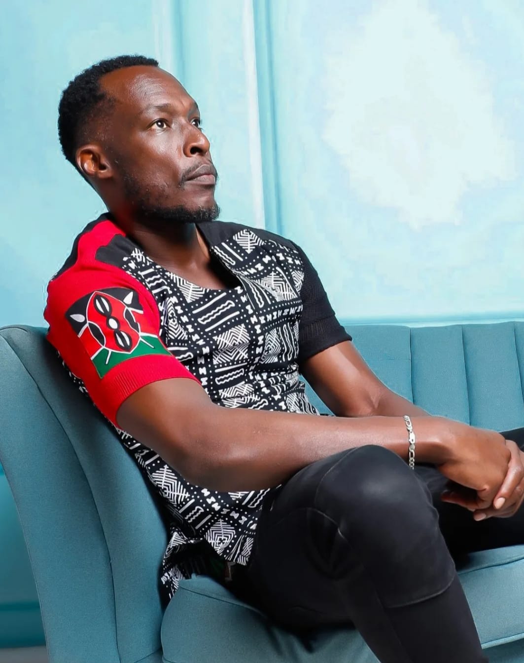 Man sitting on a teal sofa, looking to the side, wearing a patterned shirt with red and black sleeves, in a room with light blue walls.