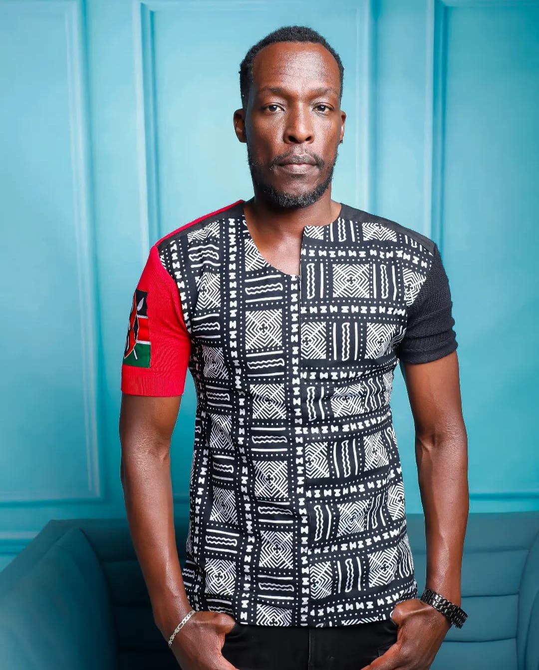 Man with short hair and beard wearing a patterned shirt, standing against a blue background.