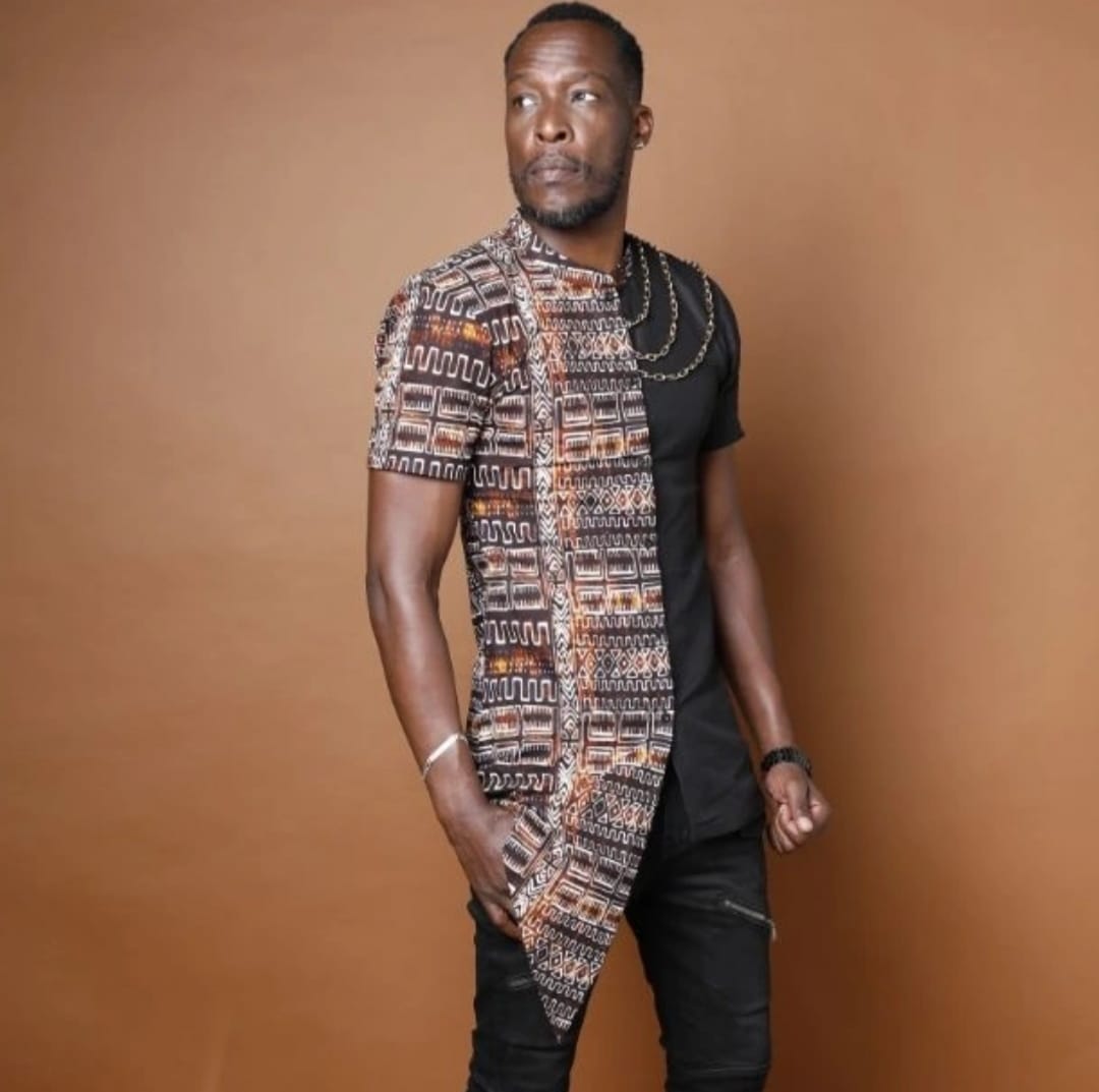Man with short hair and beard wearing a patterned shirt over a black shirt, standing against a brown background.