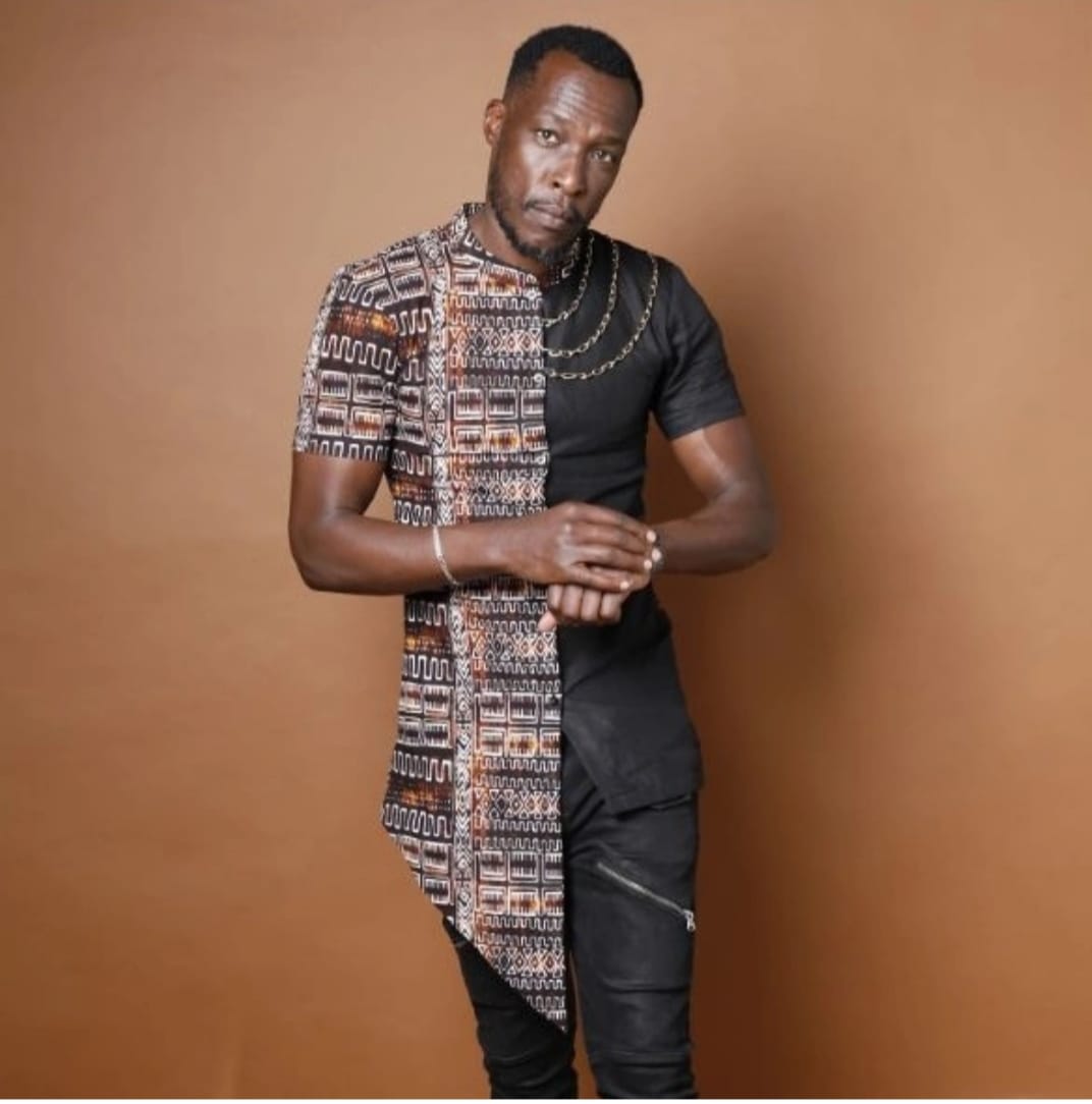 Man with short hair and dark skin wearing a black shirt and patterned fabric draped over shoulder, standing against a brown background.