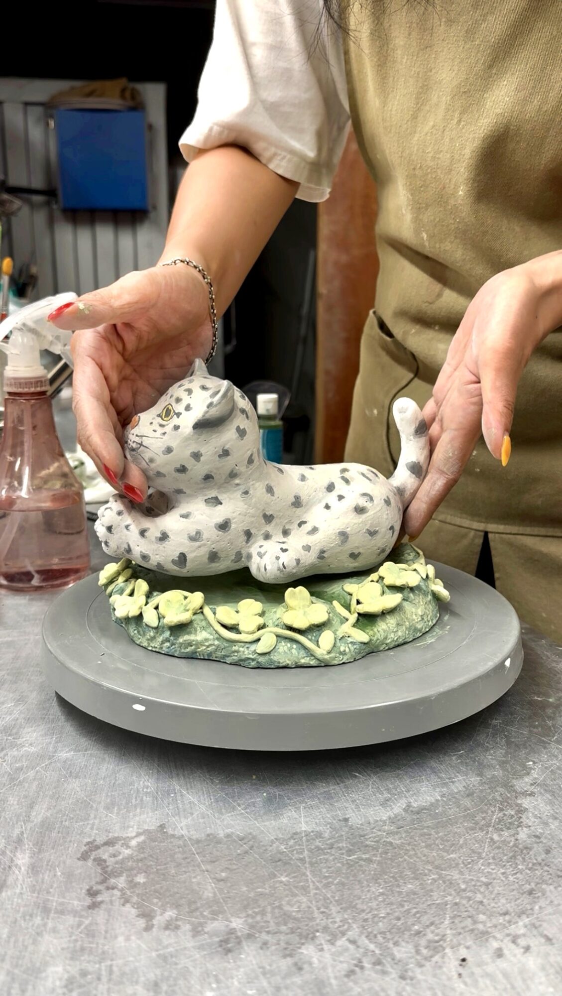 Person decorating a white, spotted animal-shaped cake with green leaves on a gray circular base.