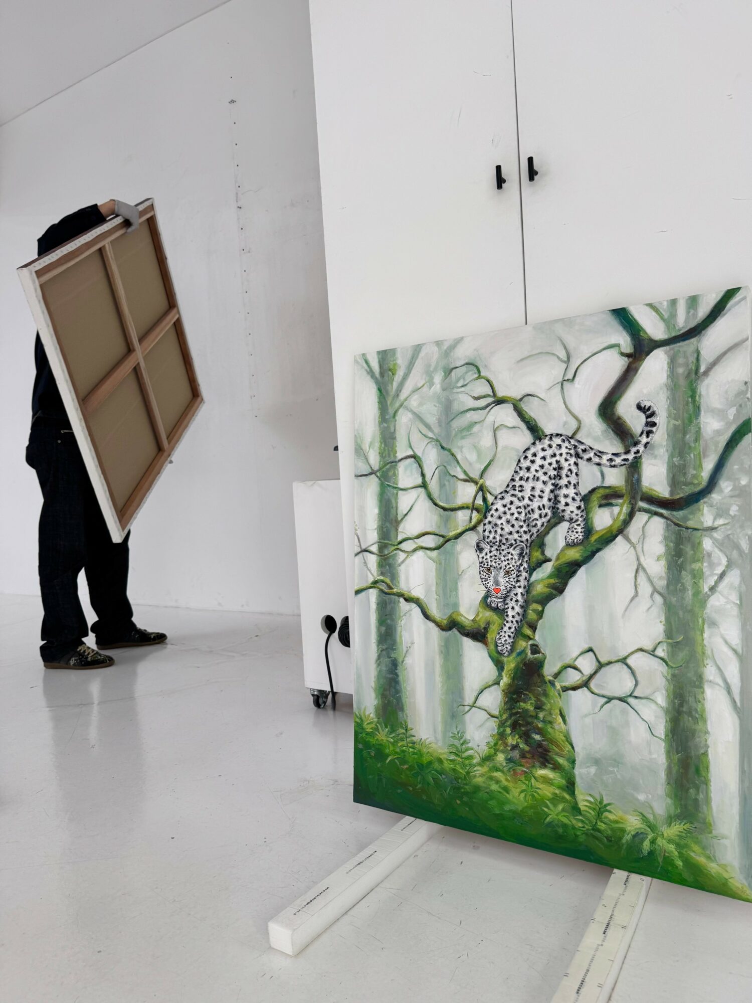 Person holding a framed artwork in a white room with a leopard painting on a tree branch.