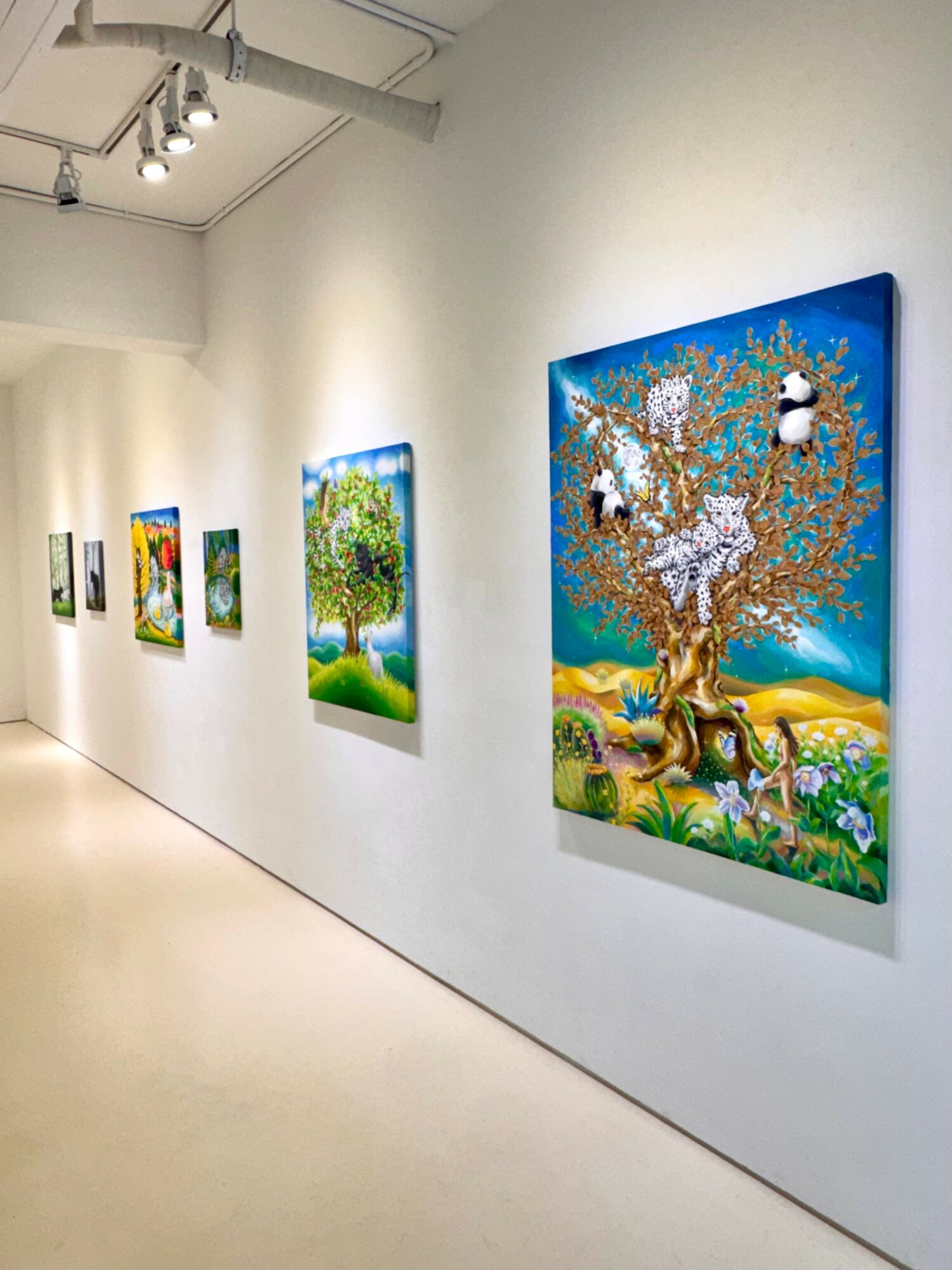 Gallery wall with colorful paintings of trees and nature scenes displayed in a bright, white room.
