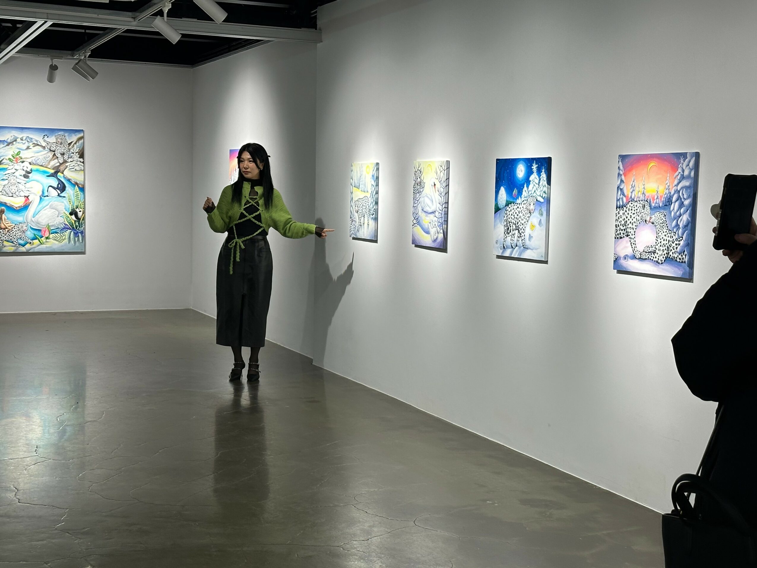 Woman in green jacket and black pants standing in art gallery, looking at colorful paintings on white wall.