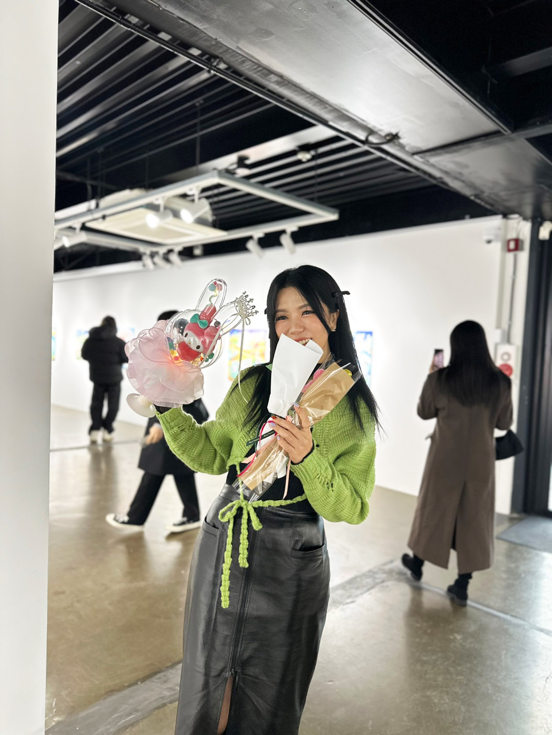 Young woman holding a colorful plush toy and a paper bag, smiling in an indoor public space with other people.