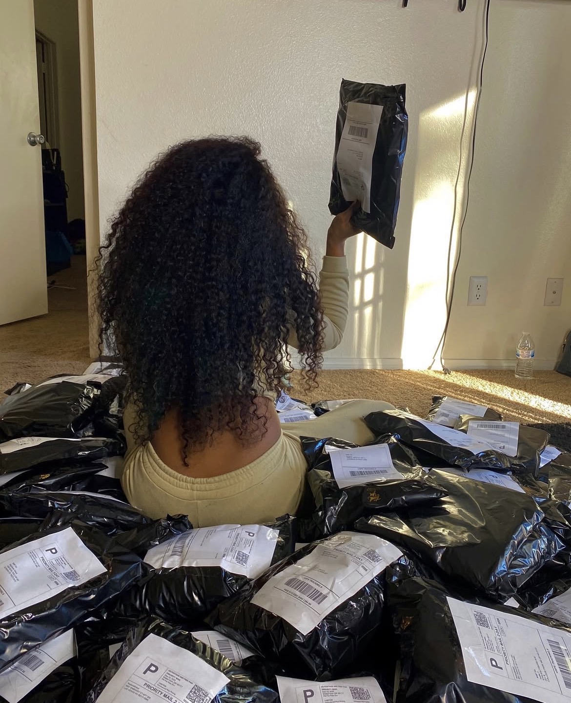 Person with curly hair sitting on a pile of black packages, holding a package up to a wall-mounted device, in a room with sunlight.