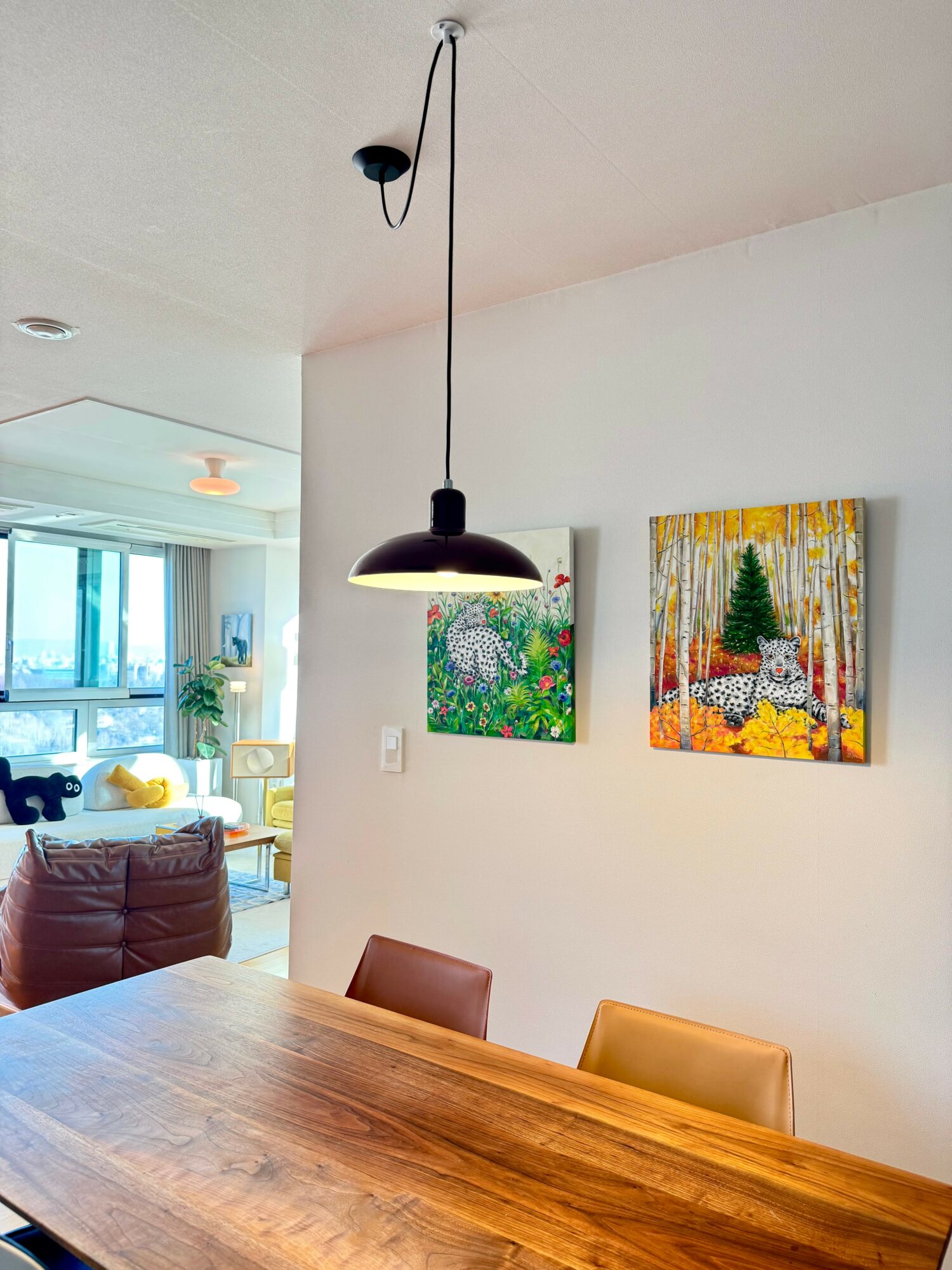 Pendant light hanging above a wooden dining table in a bright room with colorful paintings on the wall.