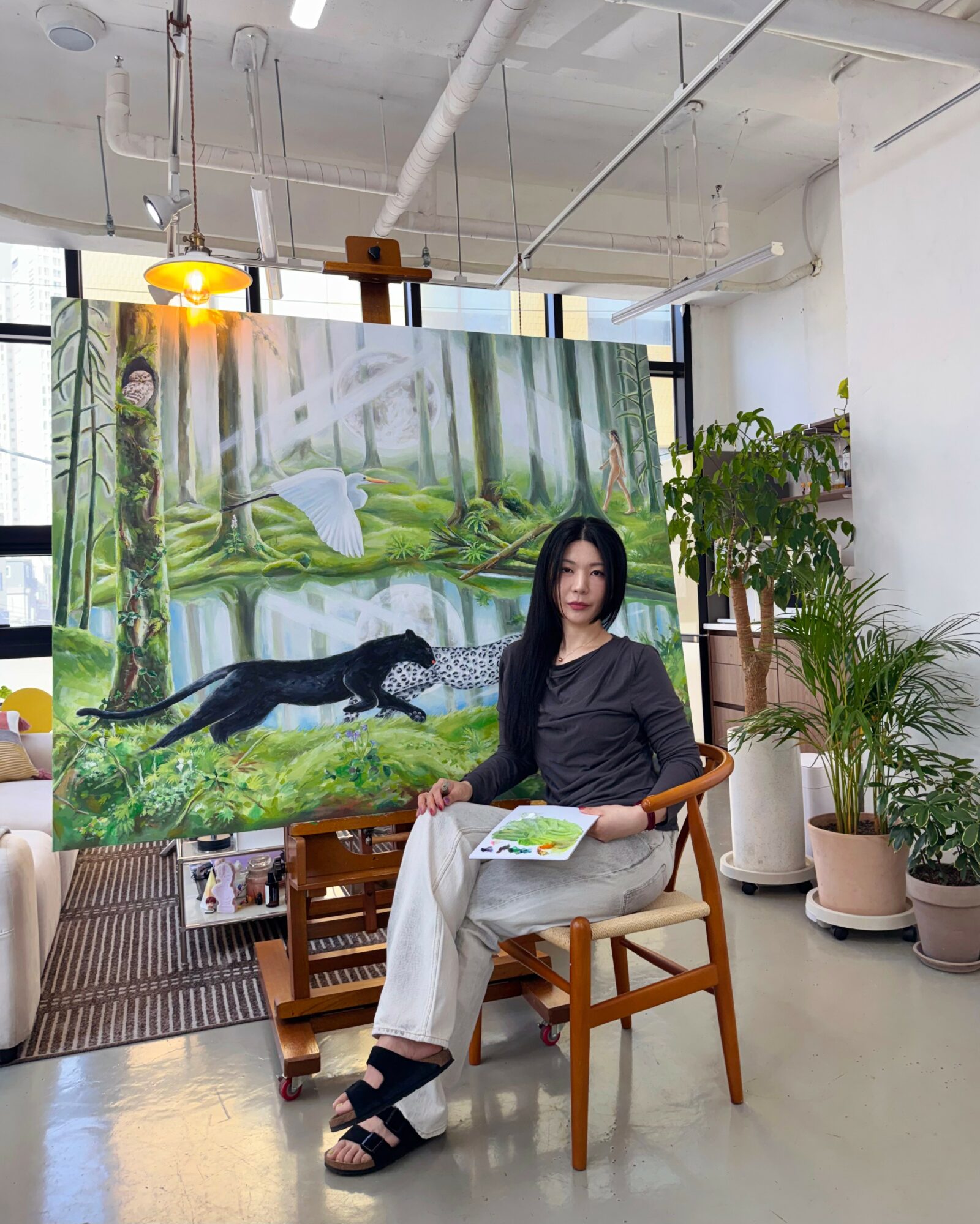 Woman sitting on a wooden chair holding a book, in front of a forest-themed painting, surrounded by potted plants.
