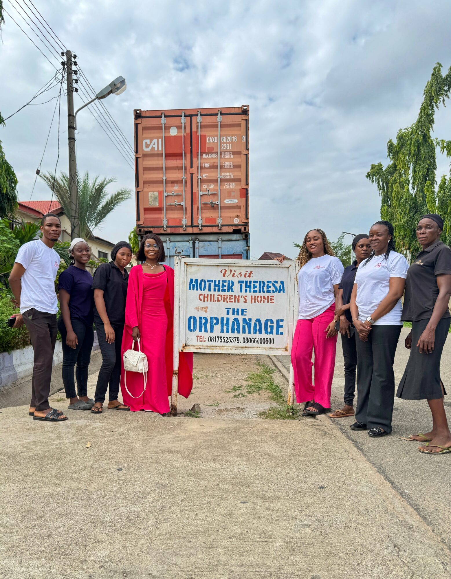 Group of nine people standing outdoors near a sign for Mother Theresa Children's Home and Orphanage, with a large container and trees in the background.