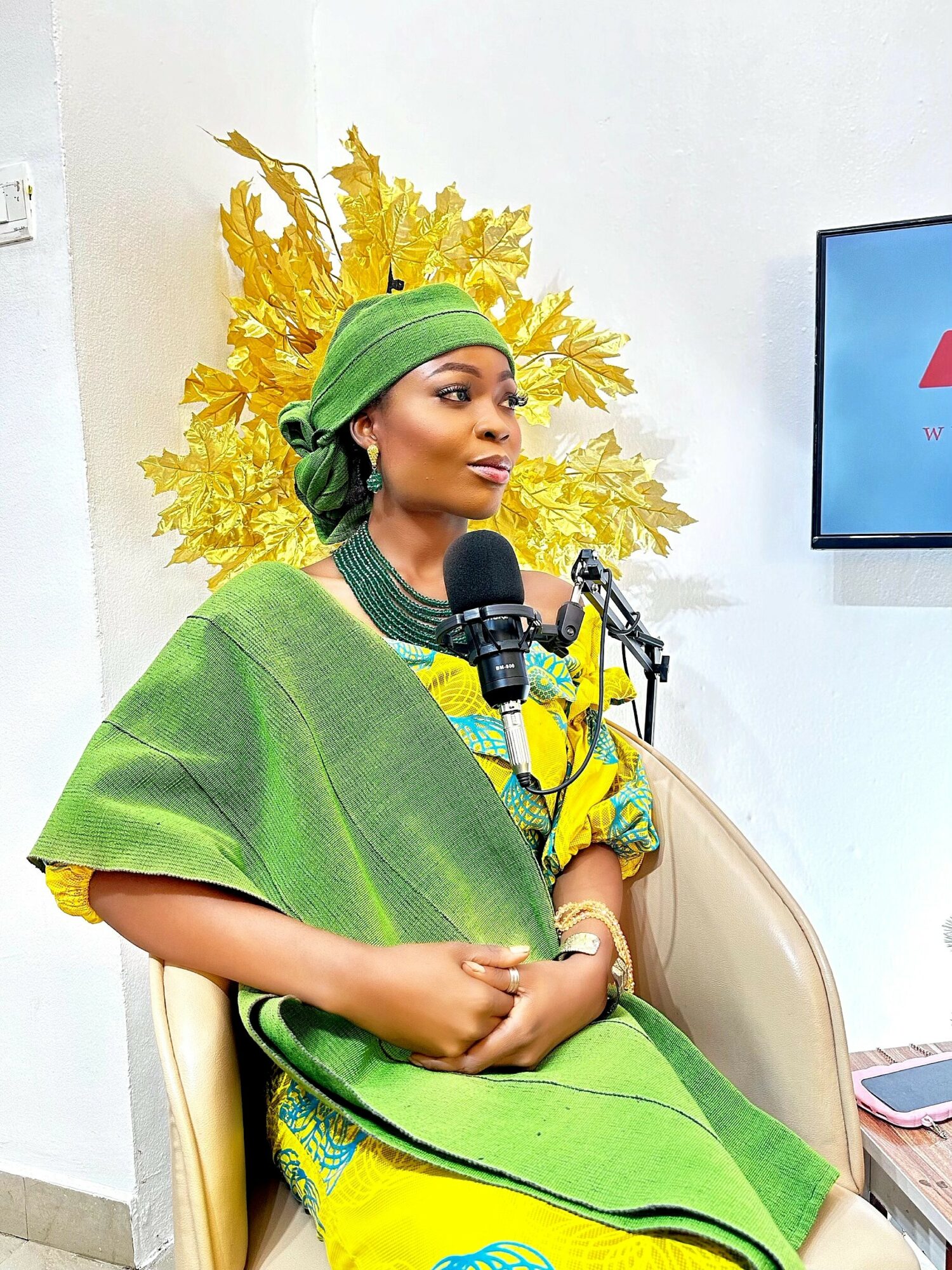 Woman in colorful traditional attire sitting with hands clasped, yellow and green background, microphone in front, TV screen on wall