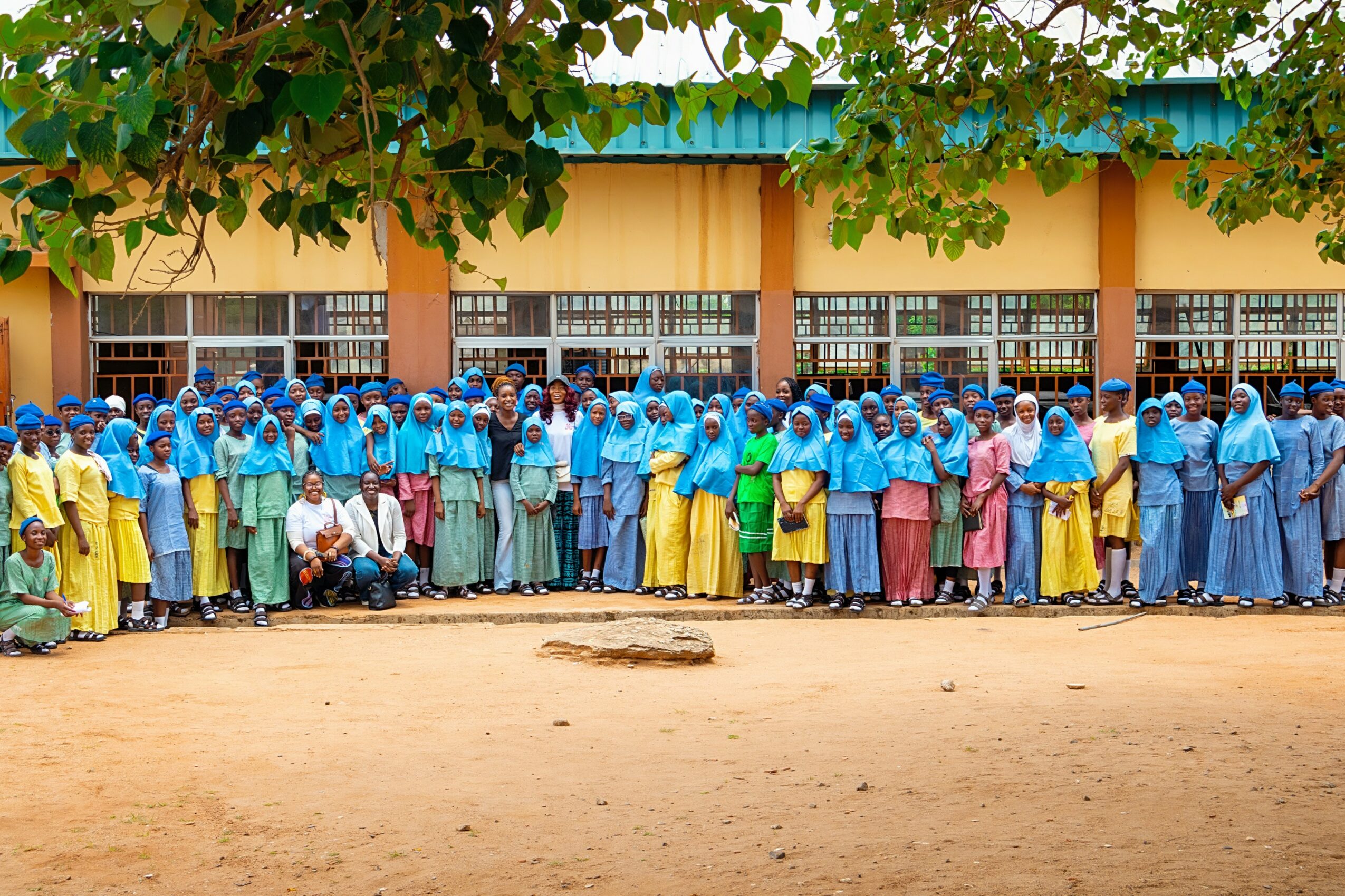 Group of people standing outside a building, some sitting, wearing colorful clothing and head coverings, under a tree.