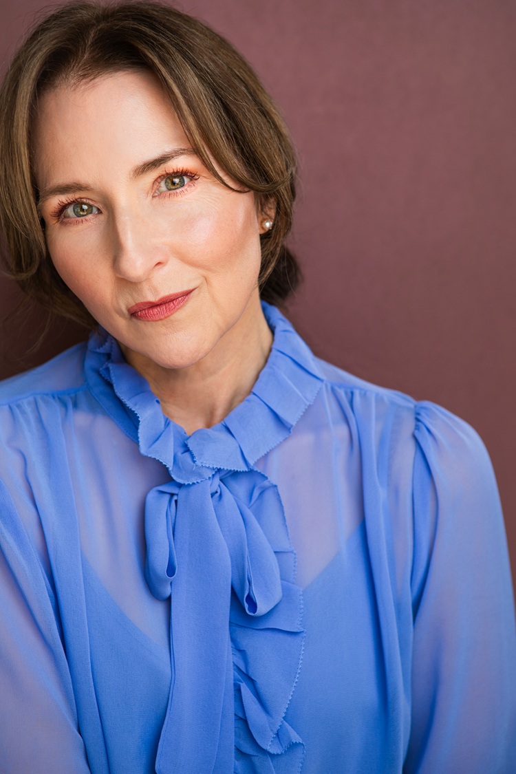 Woman with short brown hair wearing a blue blouse with a bow, smiling softly, against a plain background.
