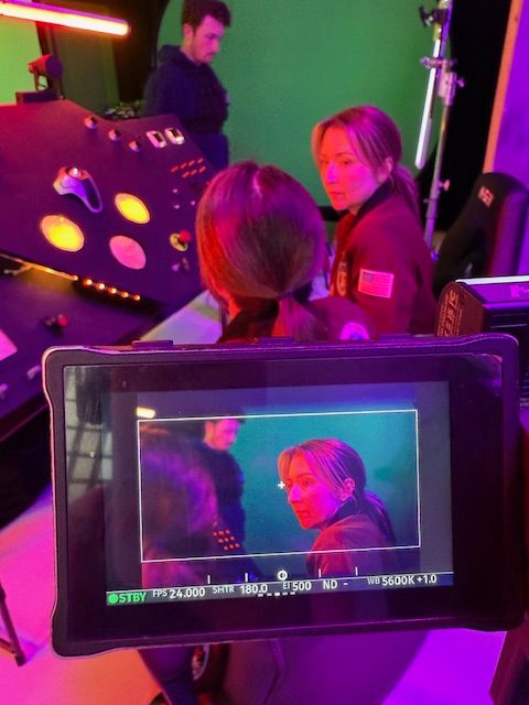 Two children with pink hair and colorful clothing are being filmed in a studio with purple lighting.