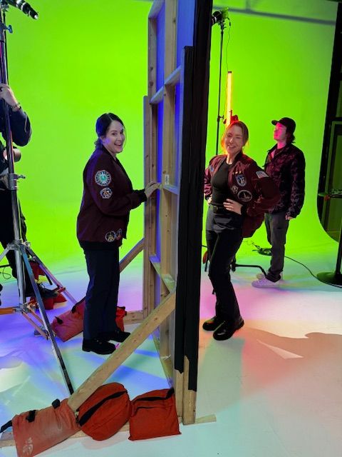 Two people in a studio with a green screen, one standing and one sitting, with equipment and a red bag nearby.