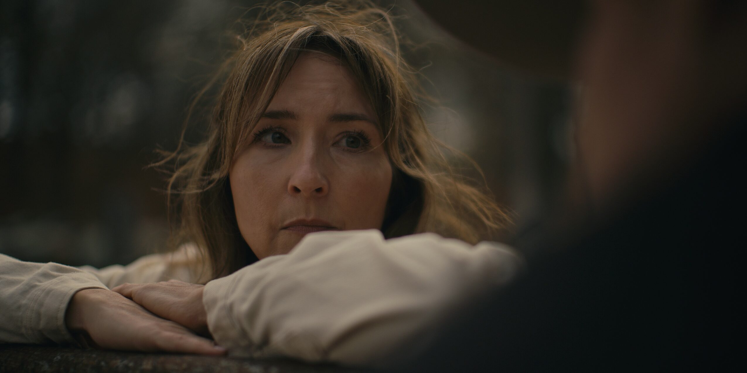 A woman rests her head on her arm, looking at a person out of focus in the foreground.