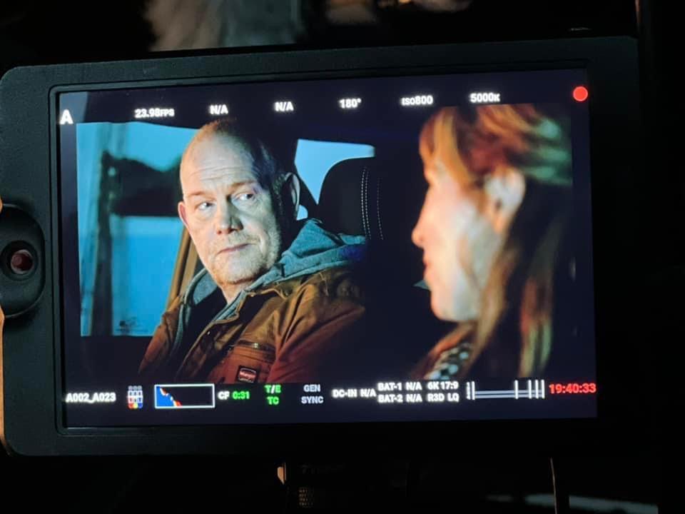 Two people sitting in a vehicle, one looking at the other, with a digital camera screen showing settings and a recording timer.