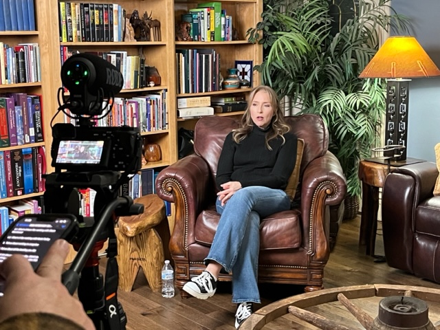 Woman sitting on a leather armchair in a room with bookshelves, plants, and a lamp, being filmed with a camera.