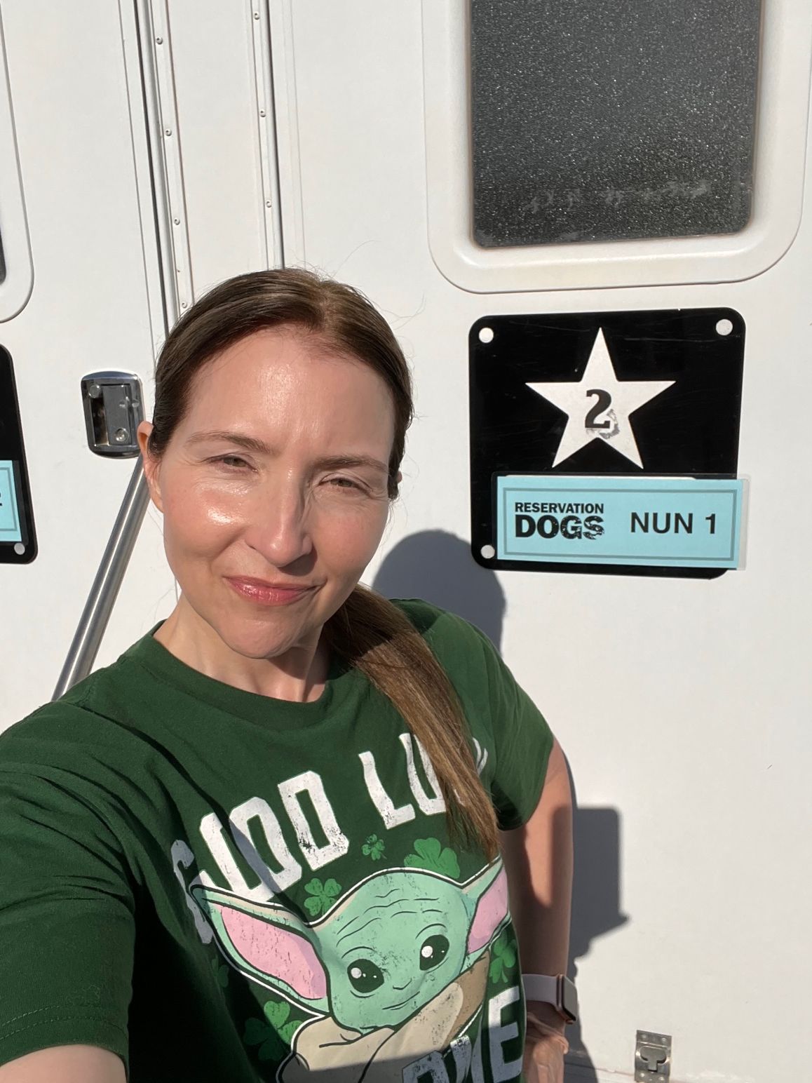 Woman with brown hair in a green shirt taking a selfie inside an elevator, with a dog sign and a star sign behind her.