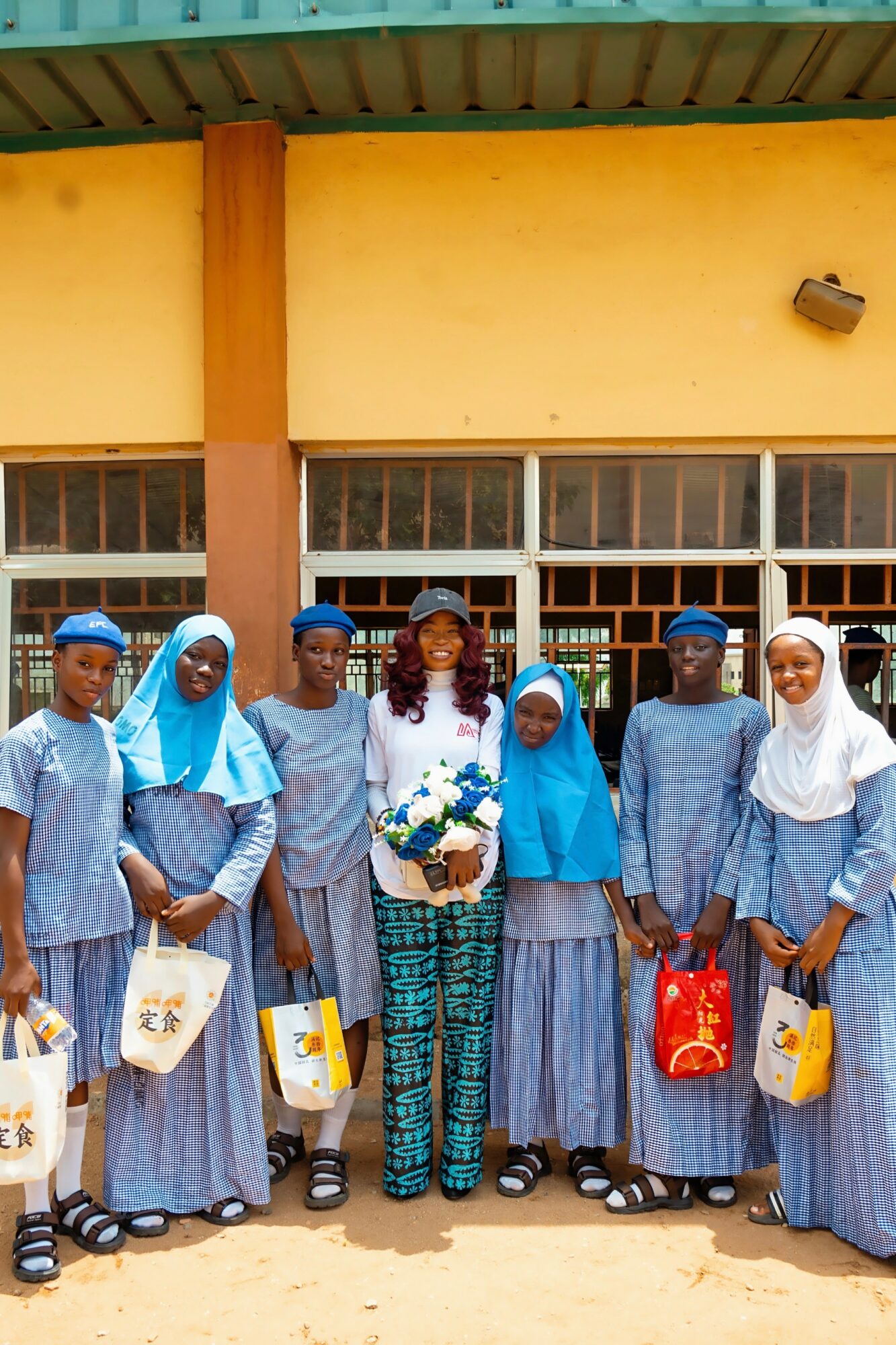 Group of seven people in blue and white uniforms standing outside a building, holding bags and flowers.