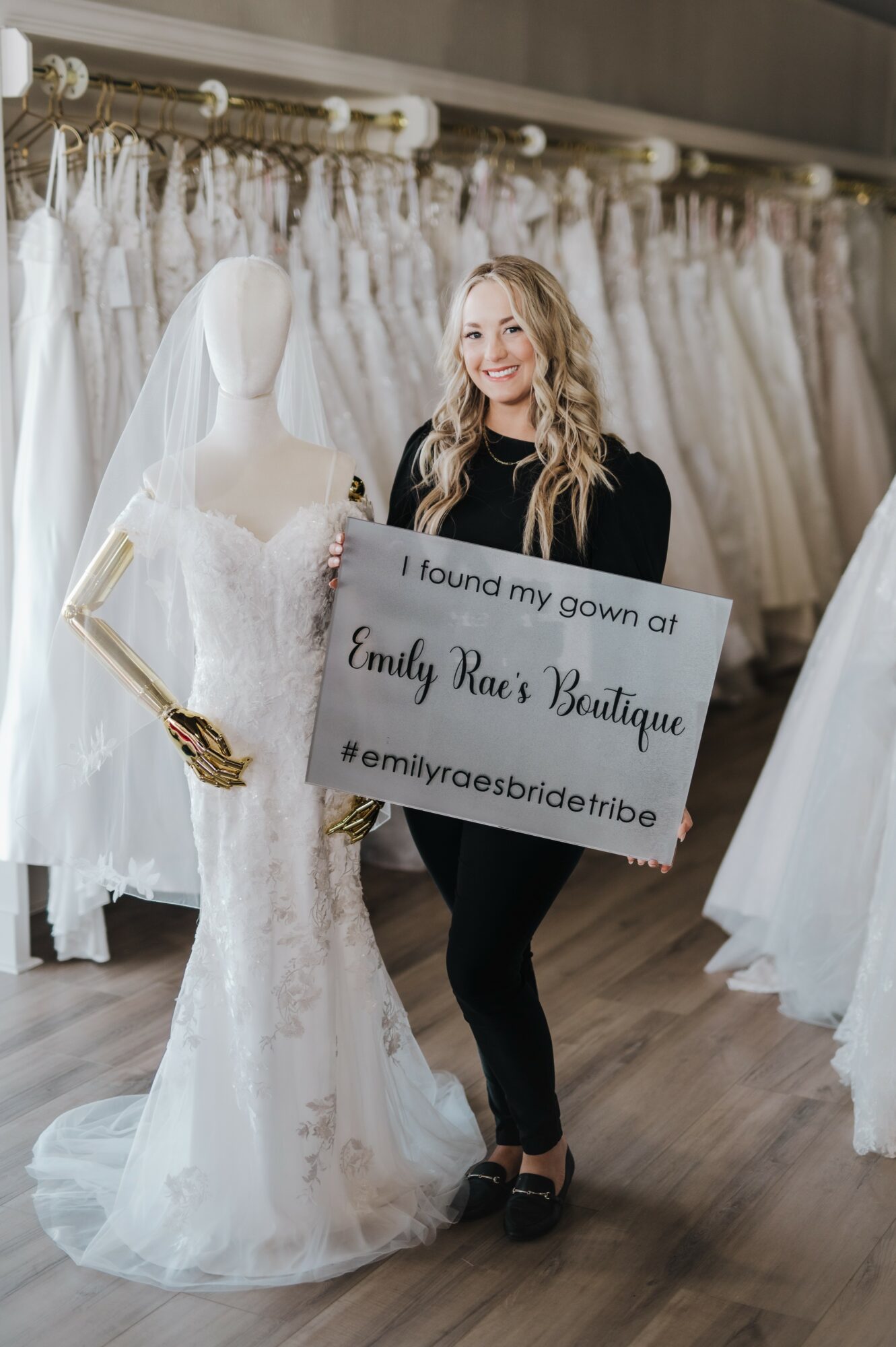 Woman with long blonde hair holding a sign in a bridal shop, surrounded by wedding dresses, standing next to a mannequin in a wedding gown.