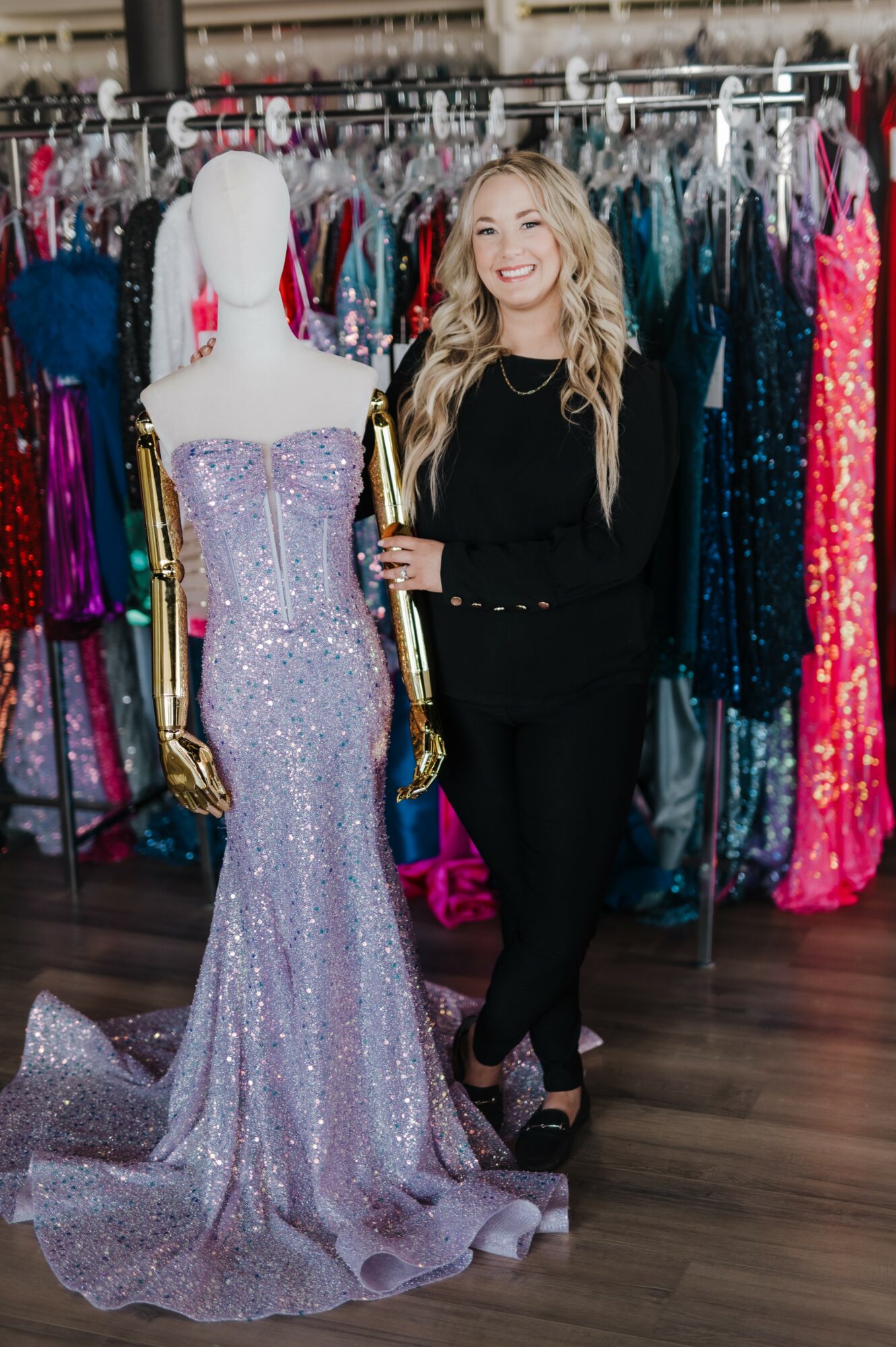 Woman with long blonde hair smiling next to a mannequin in a sparkly purple gown in a dress shop.