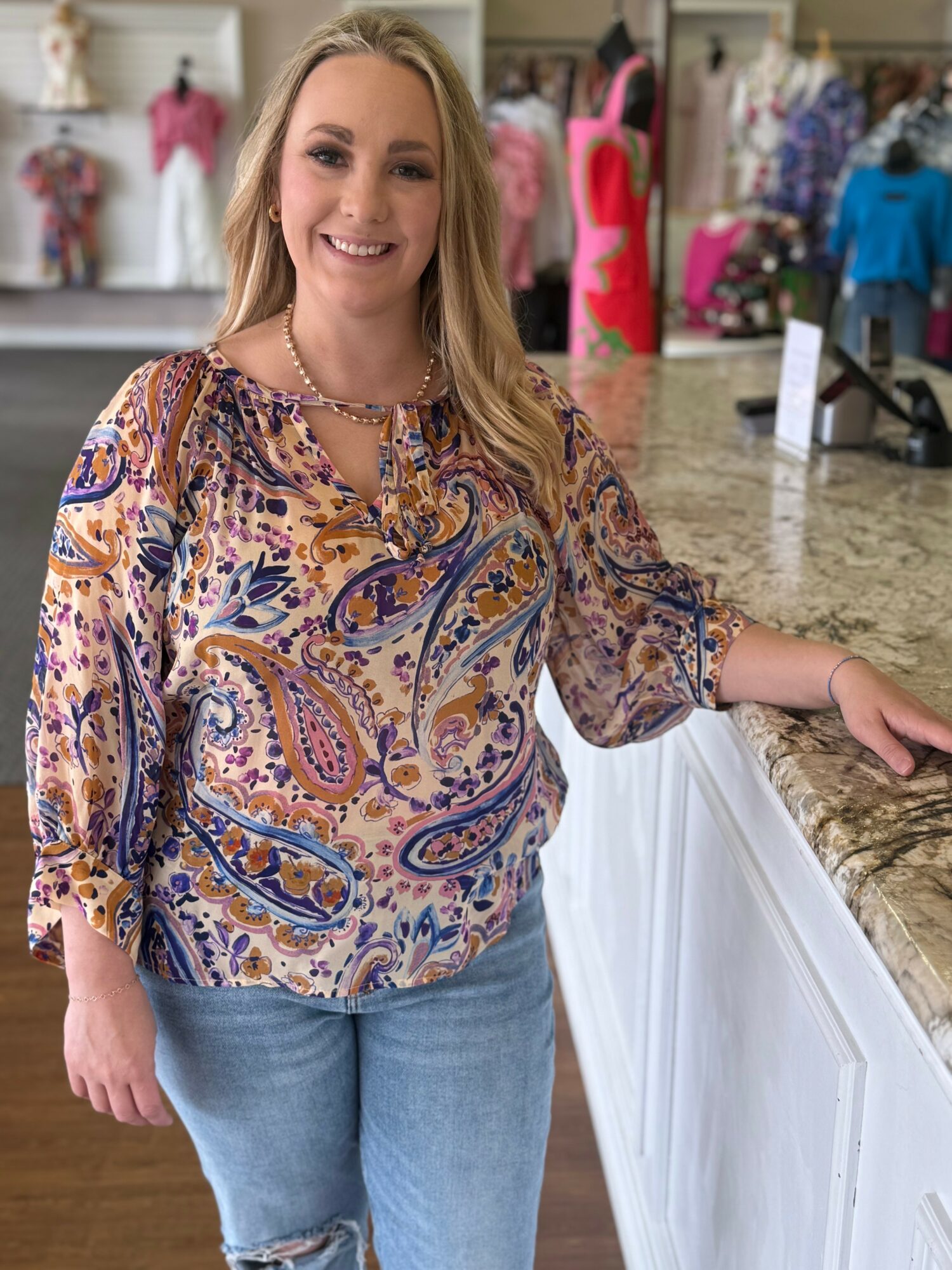 Smiling woman with long blonde hair standing at a store counter, wearing a colorful paisley blouse and jeans.