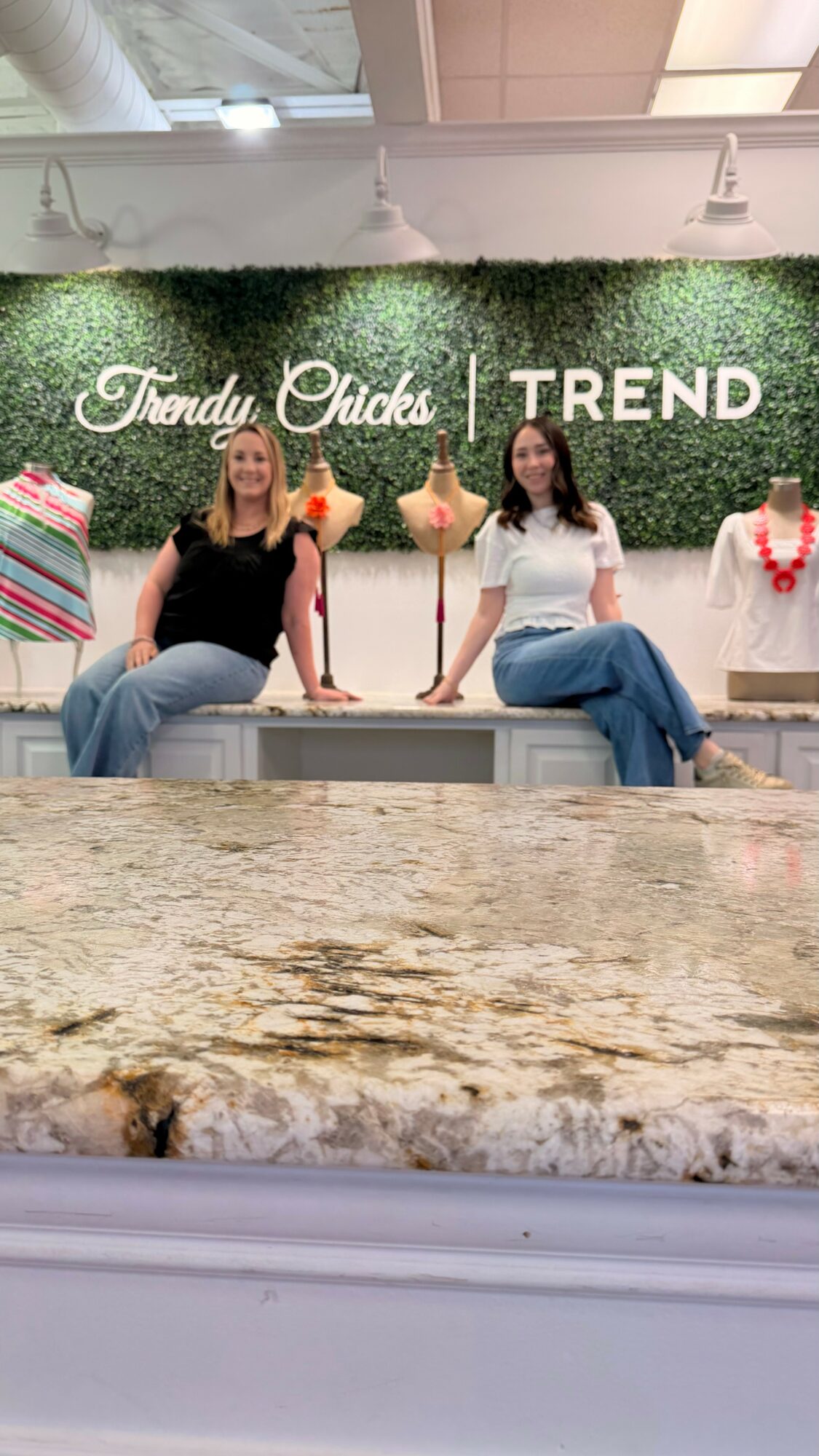 Two women sit on a counter in front of a green wall with signs reading 