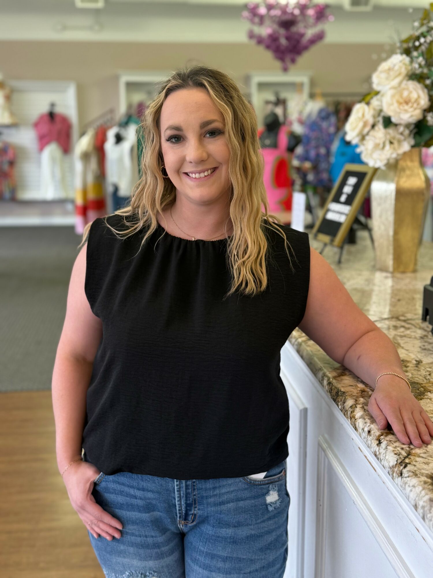 Woman with wavy blonde hair smiling, wearing a black sleeveless top and blue jeans, standing in a clothing store.